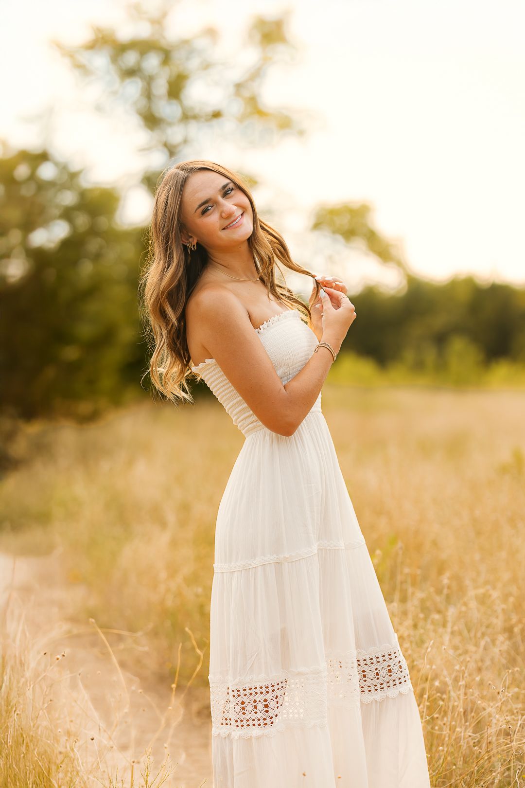 A person in a white lace dress poses joyfully in a golden sunlit field during a summer photoshoot.