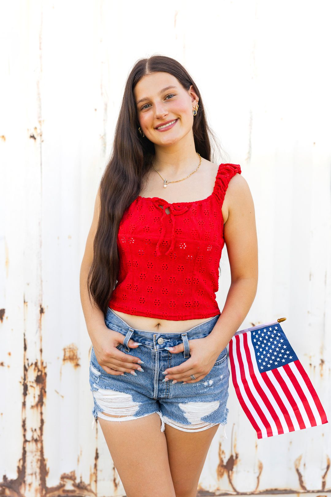 Smiling young woman in red crop top and denim shorts holding an American flag against a white wall.