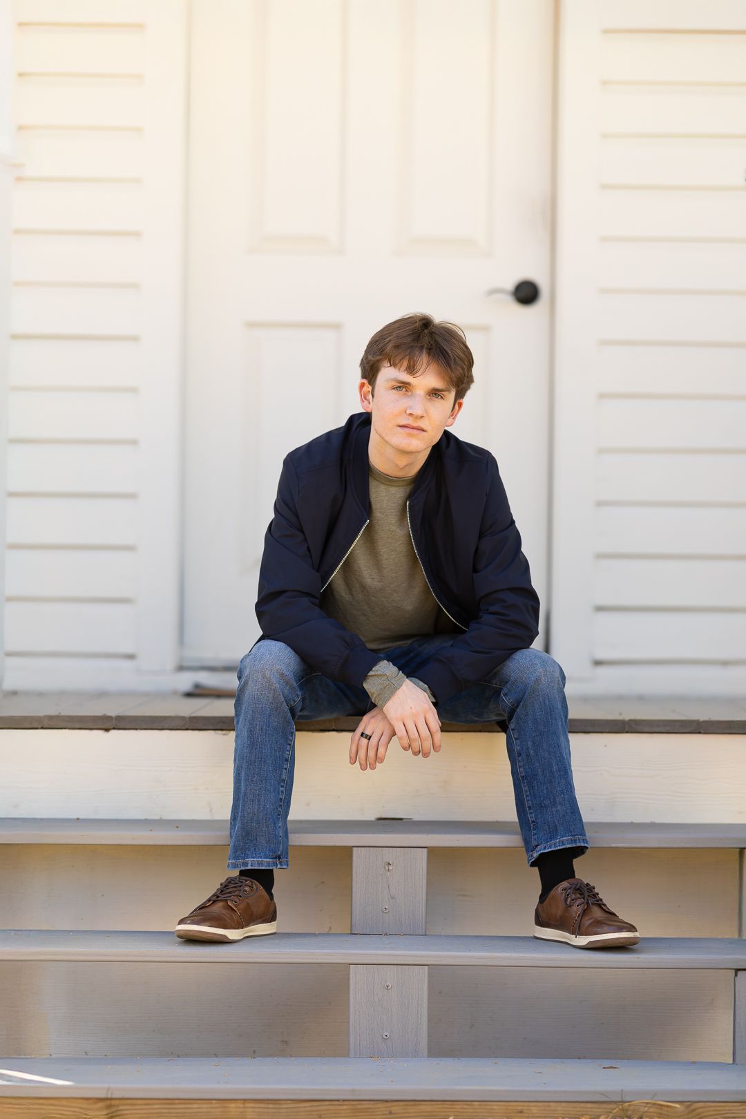 A person in a navy blazer and jeans sits on a white ledge in front of a white garage door.