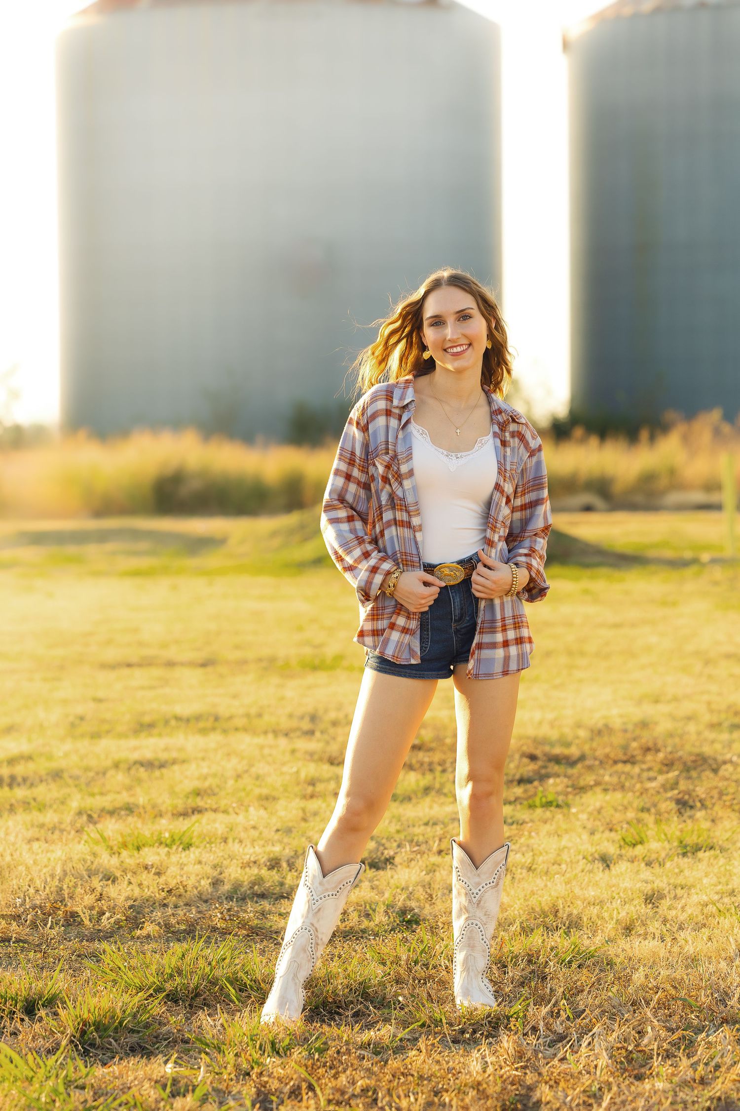 A person in a plaid shirt and white cowboy boots poses in a golden field at sunset with grain silos in the background.