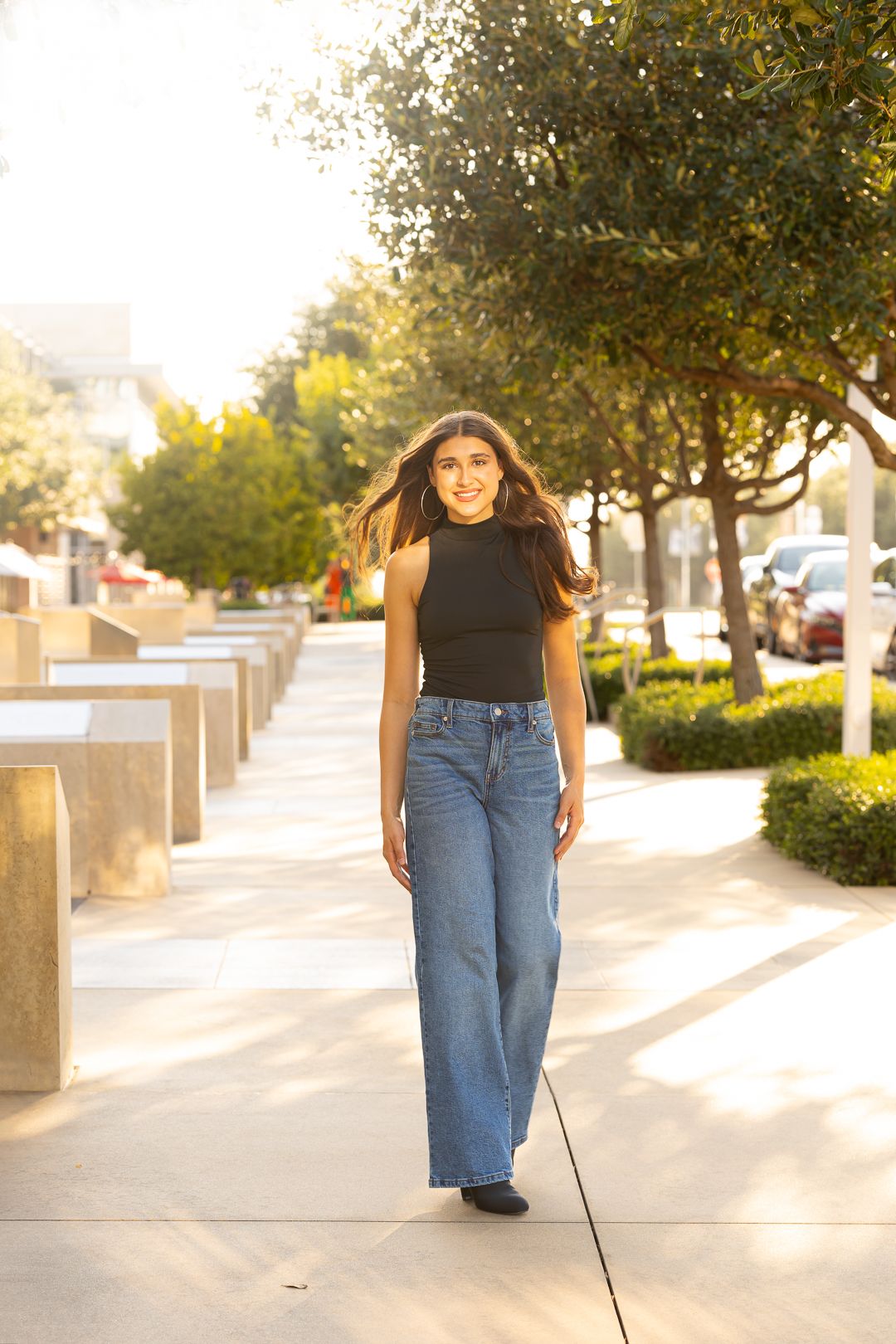 A person in a black top and blue jeans walks along a sunlit sidewalk lined with trees on a bright afternoon.