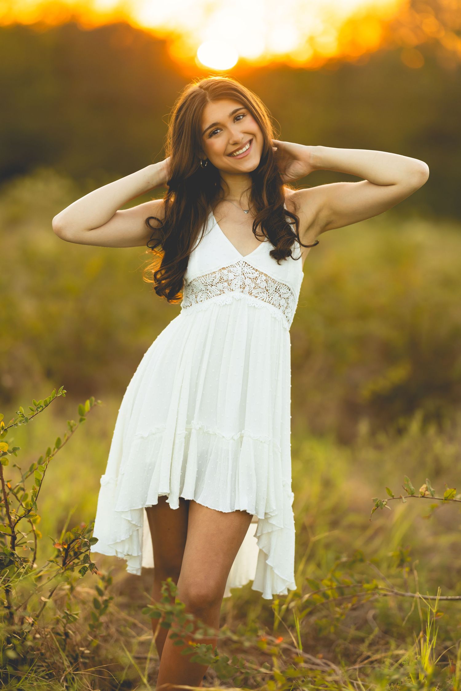 A person in a flowing white sundress poses playfully in a golden field at sunset.