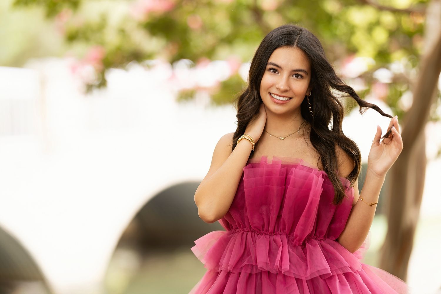 A person in a flowing burgundy dress poses outdoors in a natural setting with sunlit trees in the background.