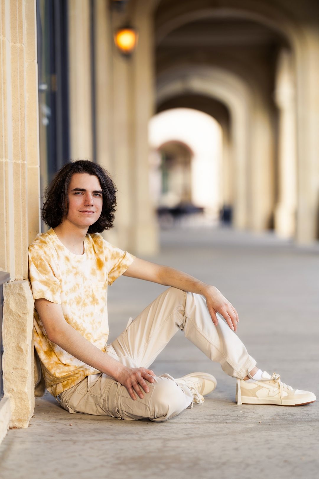 Person in yellow tie-dye shirt and khaki pants sits casually against stone archway in outdoor corridor.