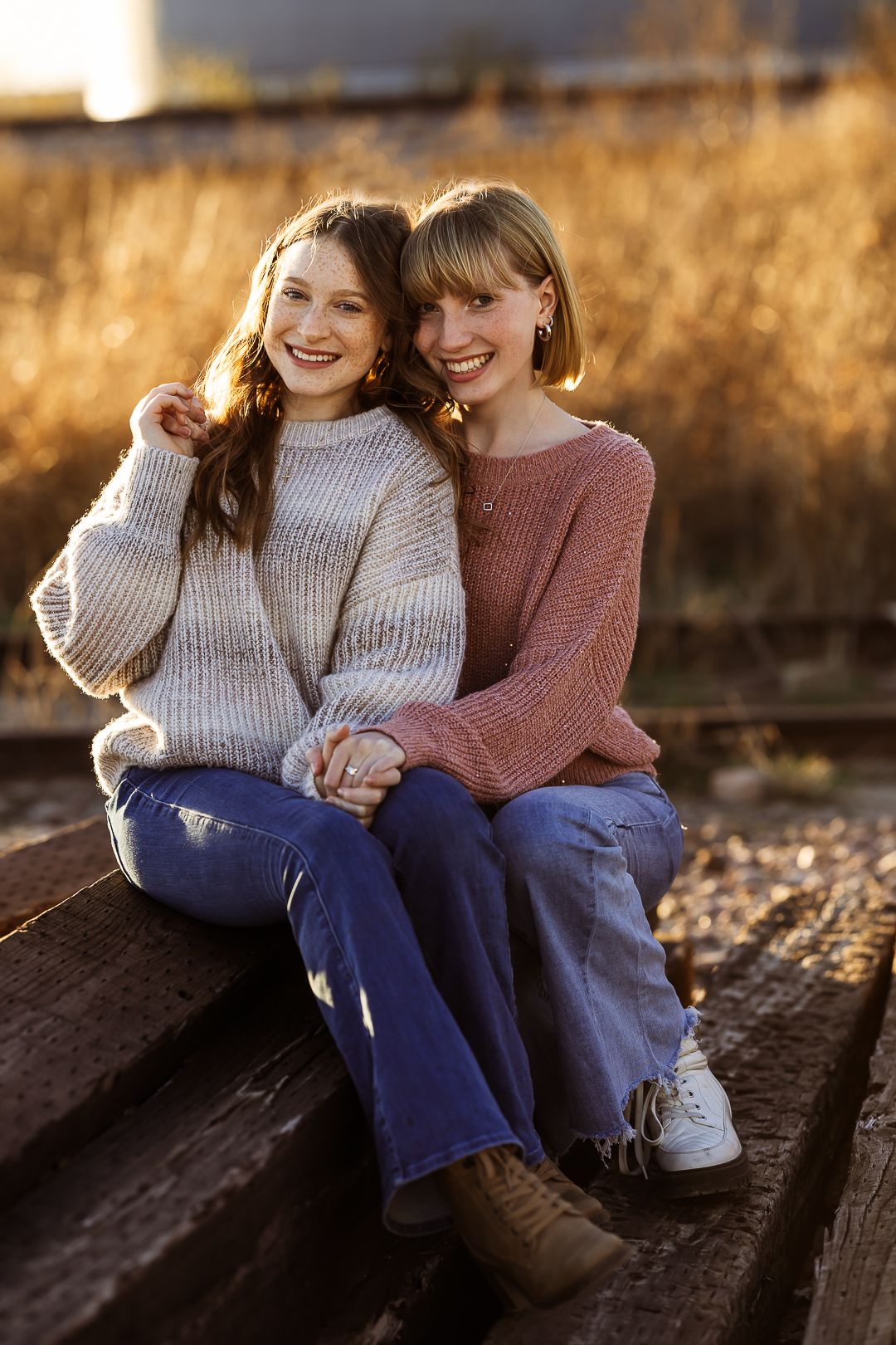 Two people in cozy sweaters sit close together on wooden steps during golden hour autumn light.