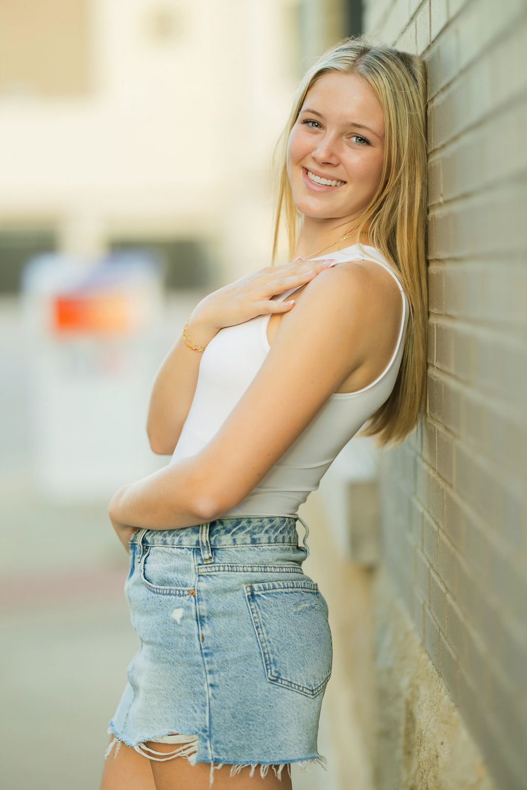 A person in a white top and denim shorts poses against a light colored wall.