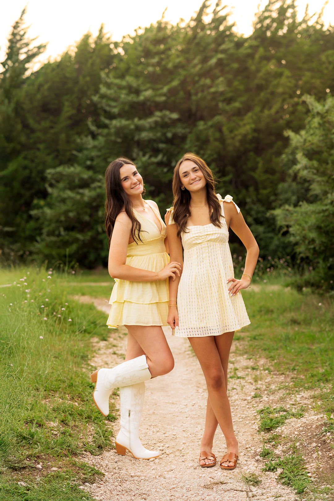 Two young women in matching yellow mini dresses pose on a dirt path surrounded by lush green trees.