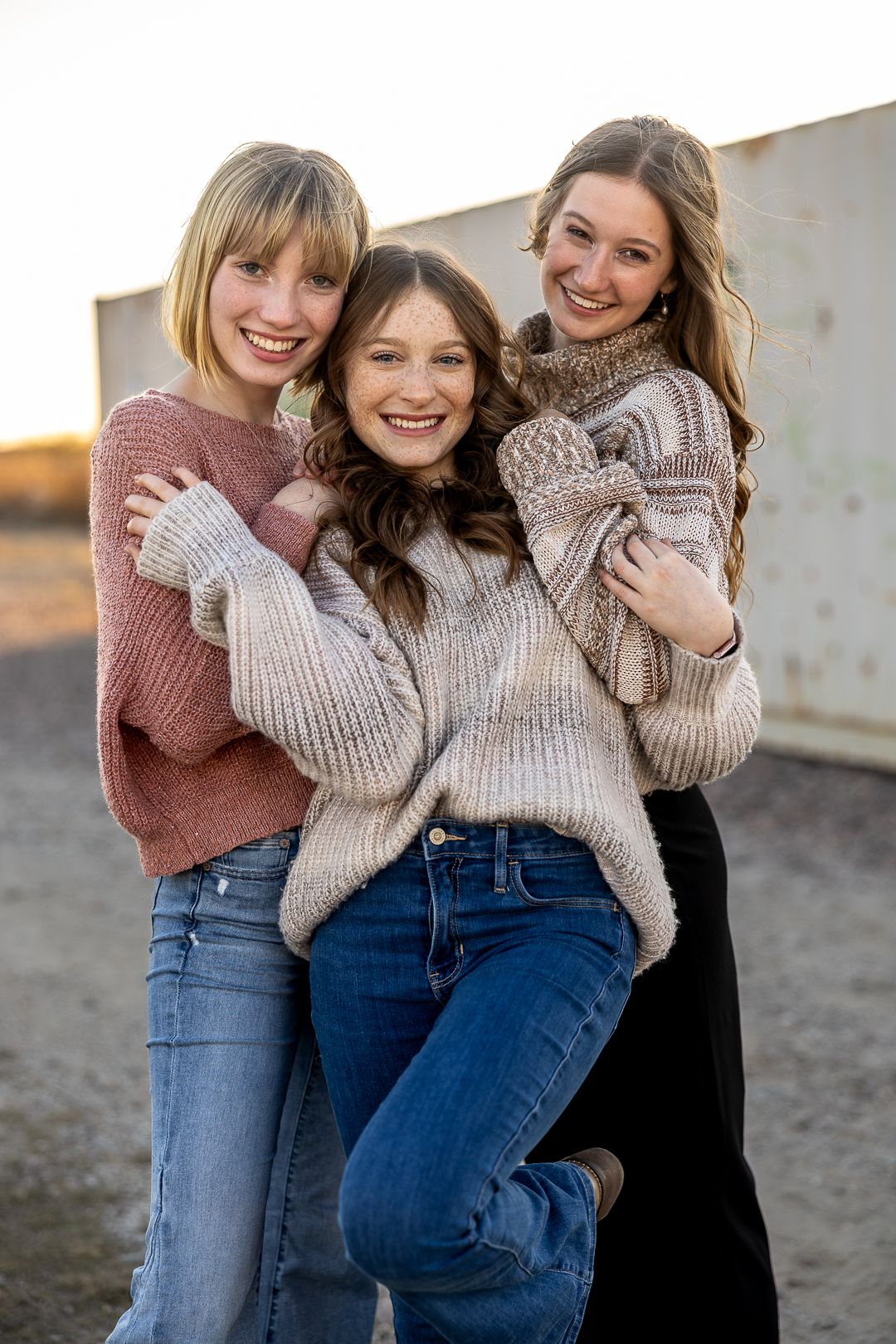 Three people wearing cozy sweaters and jeans embrace and laugh together during an outdoor photoshoot.