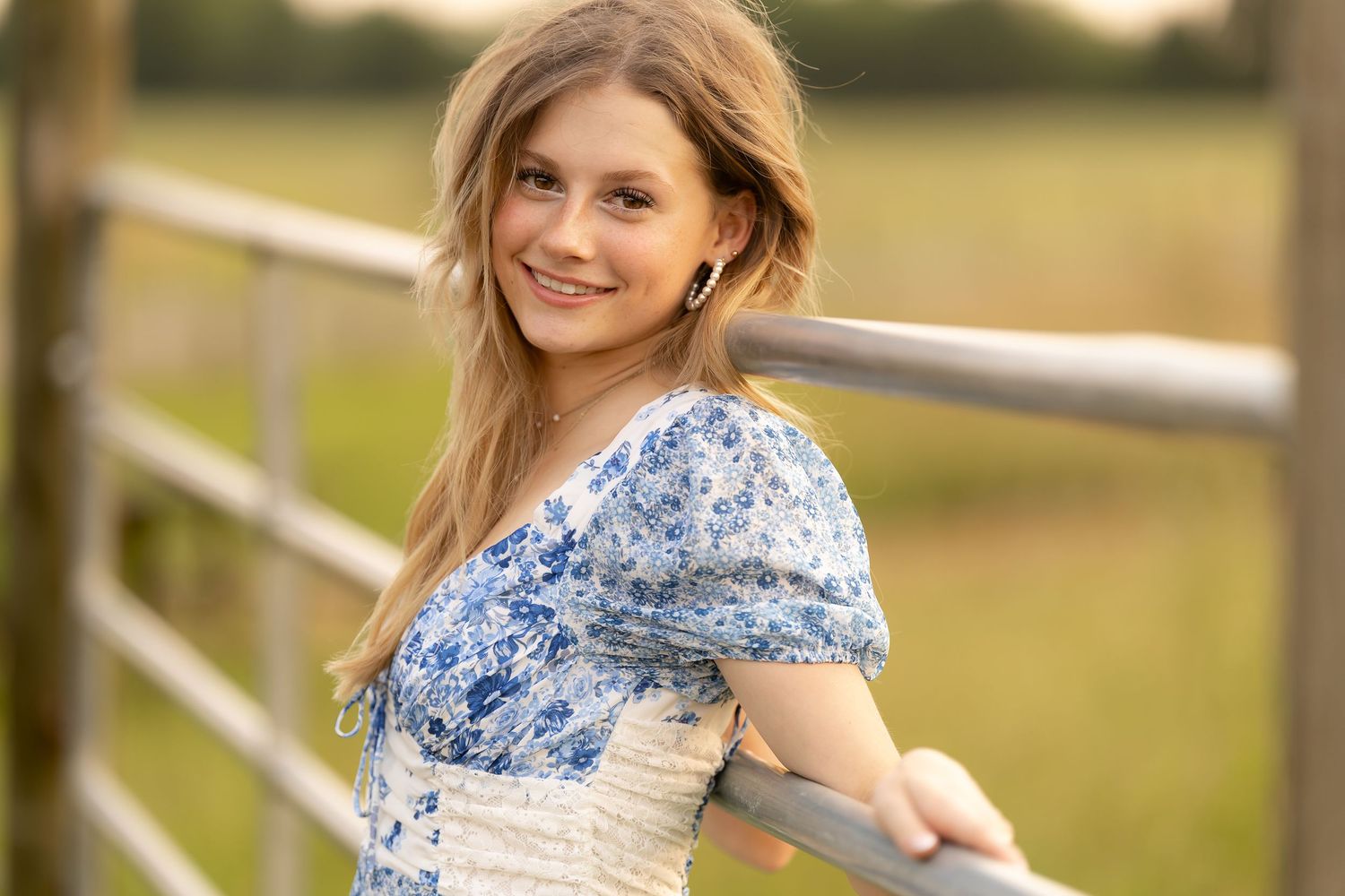 A person in a blue floral dress leans against a white fence rail in a sunny outdoor setting.
