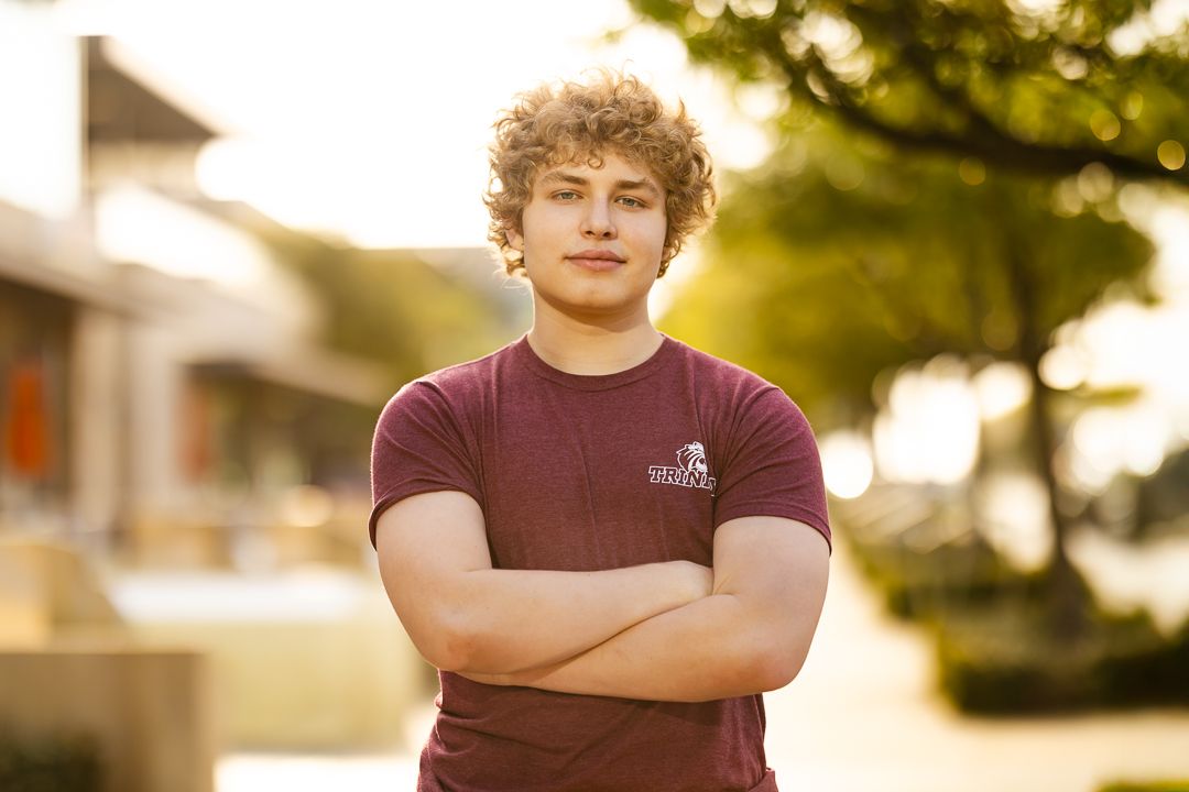 A person in a burgundy t-shirt stands with crossed arms in warm sunset lighting outdoors.