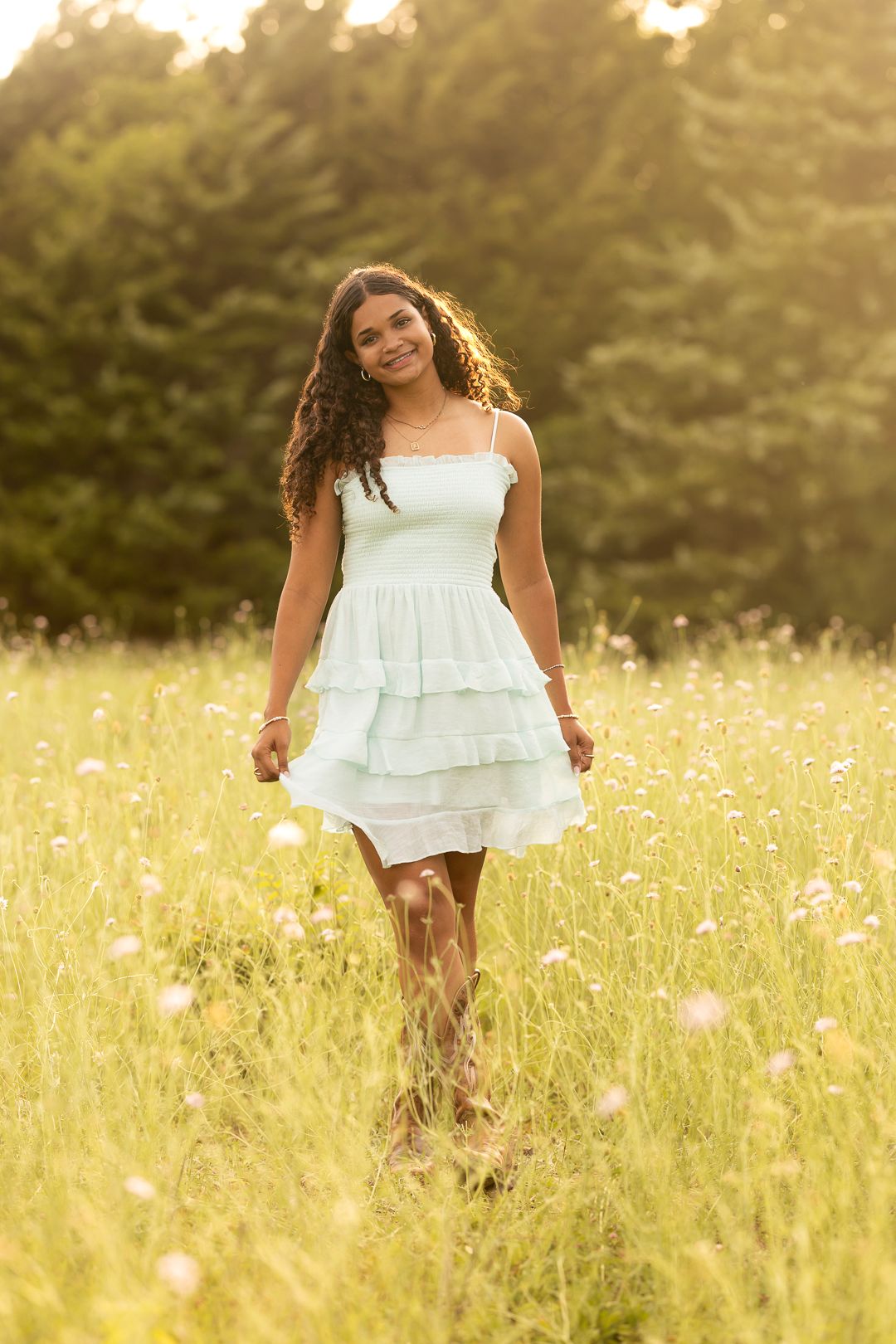 Young woman in white ruffled dress walking through sunny wildflower meadow with green trees in background.