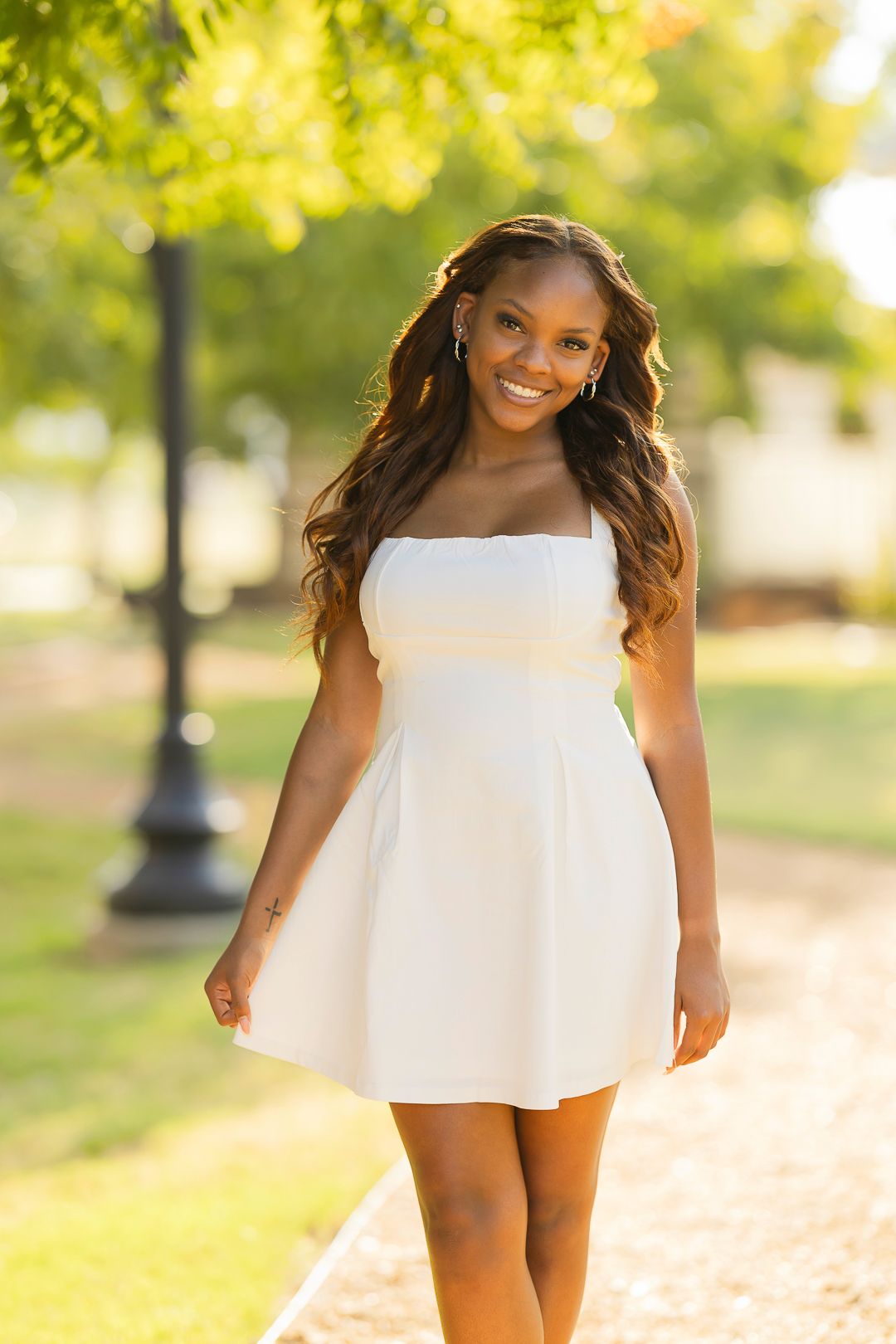 Someone in a flowing white dress stands on a sunlit path under green trees.