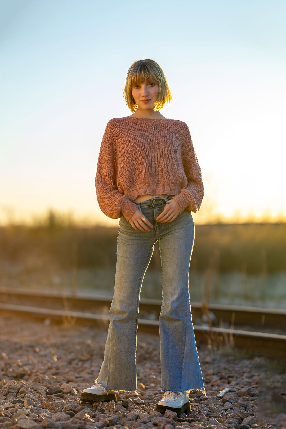 Person wearing brown sweater and jeans poses on railroad tracks at dusk.