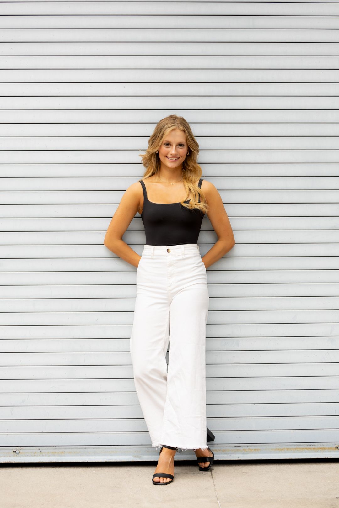 Fashion model in black top and white pants poses against white horizontal siding.