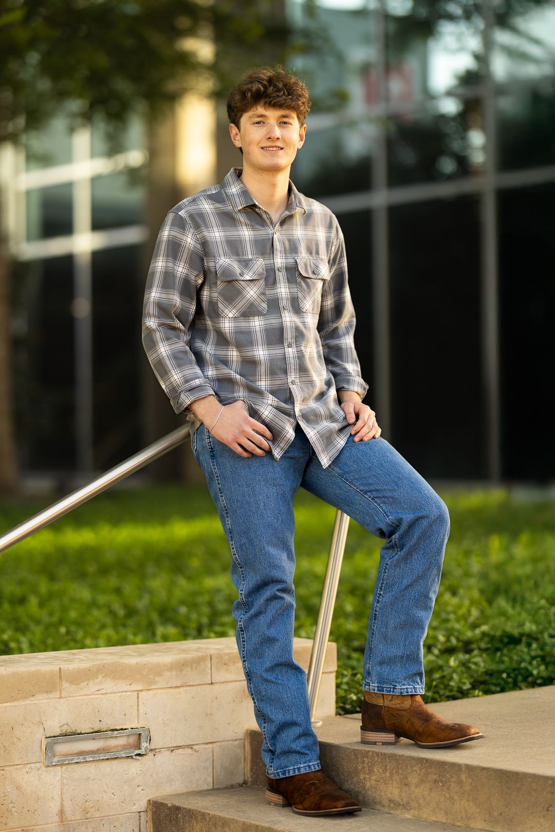 A person in a plaid shirt and jeans sits casually on outdoor concrete steps in a garden setting.
