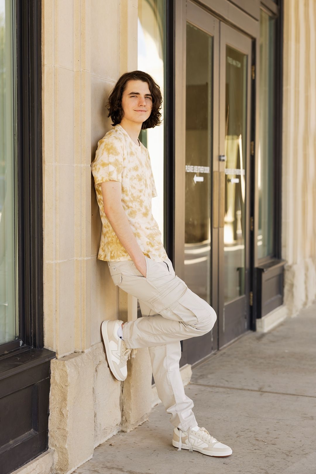A person in a floral top and white pants poses against a beige wall in a modern building corridor.