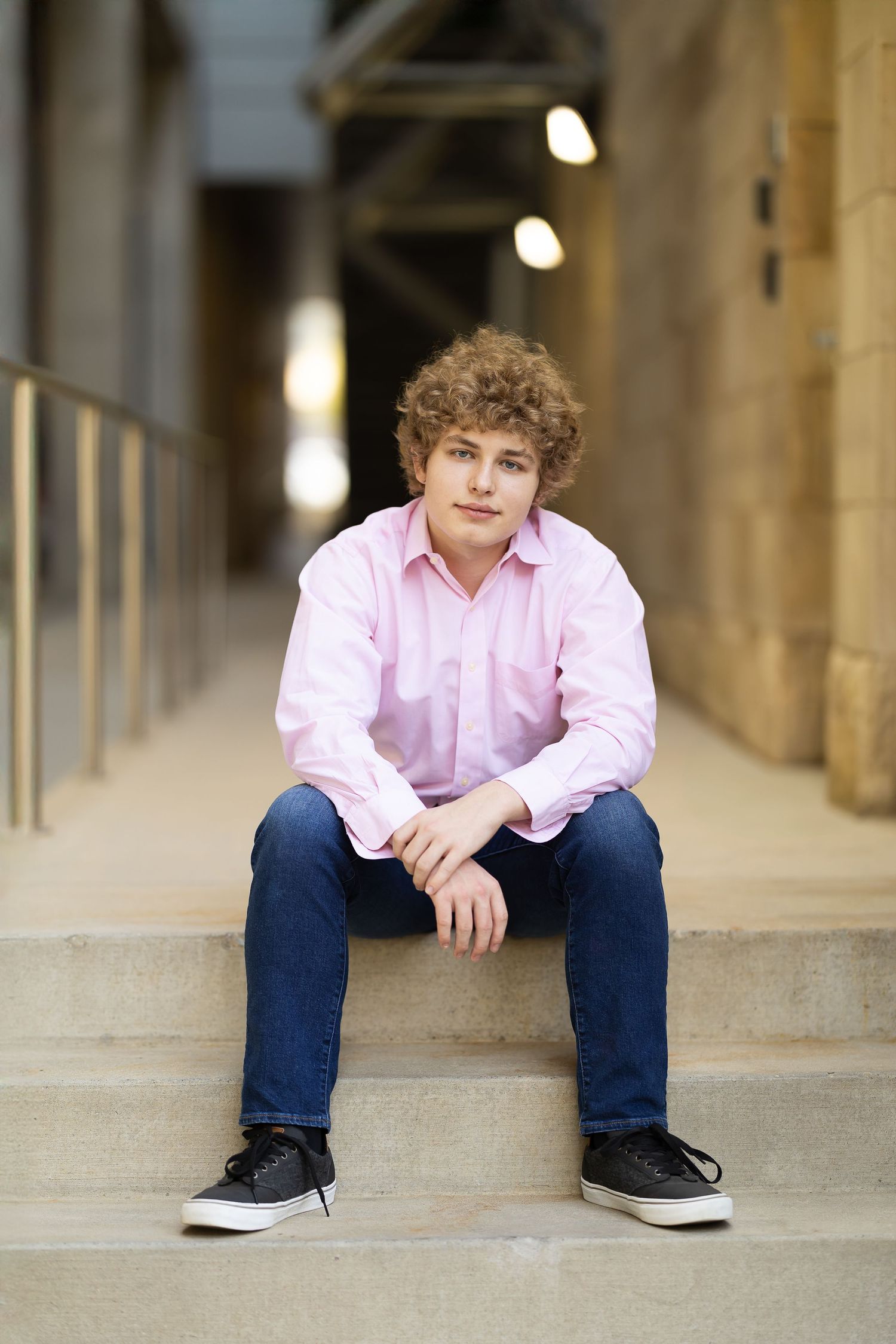 Young person in pink button-down shirt and jeans sits casually on concrete steps in an architectural setting.