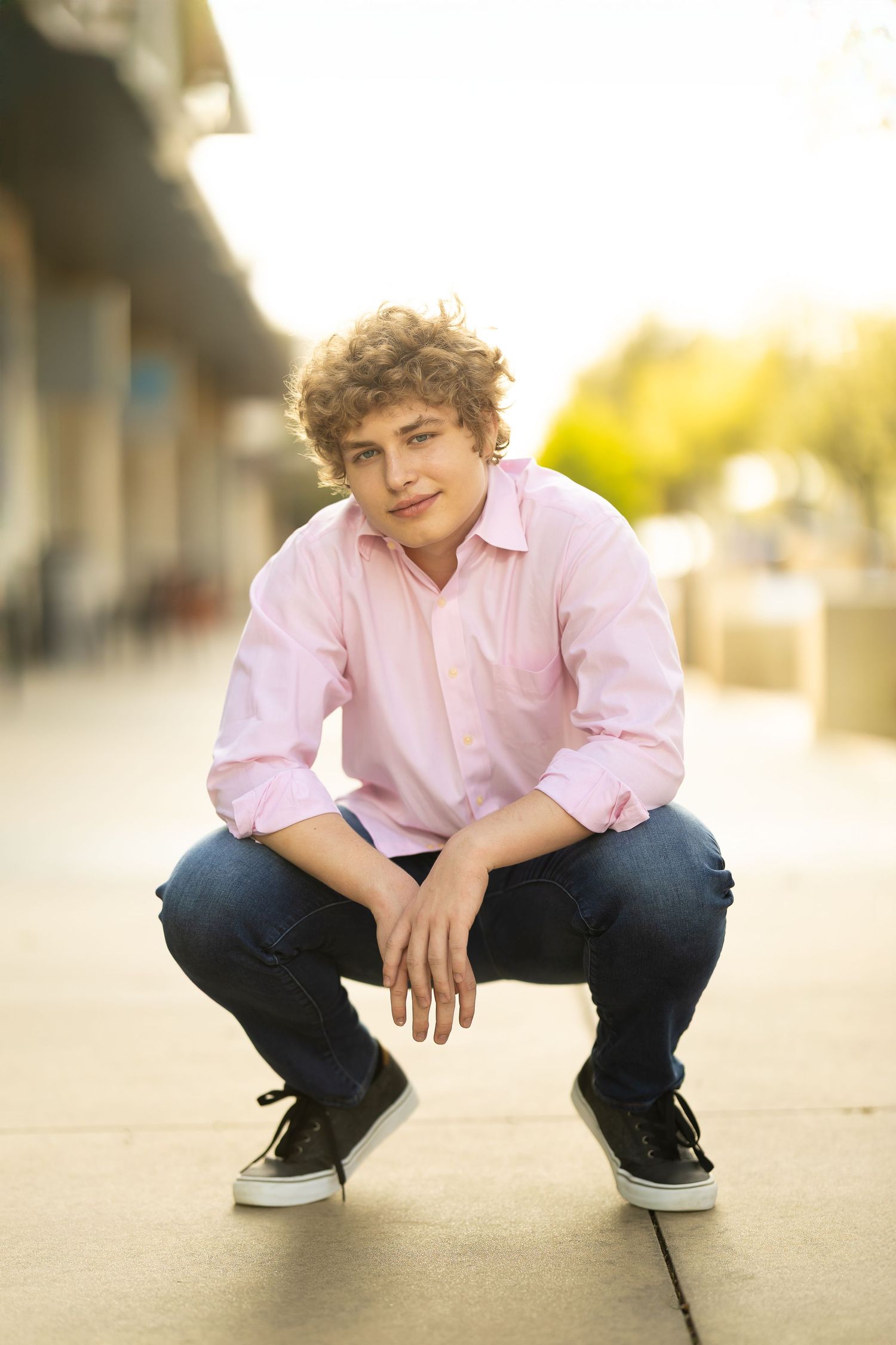 A person in a pink button-down shirt and jeans crouches down in an outdoor setting during golden hour lighting.