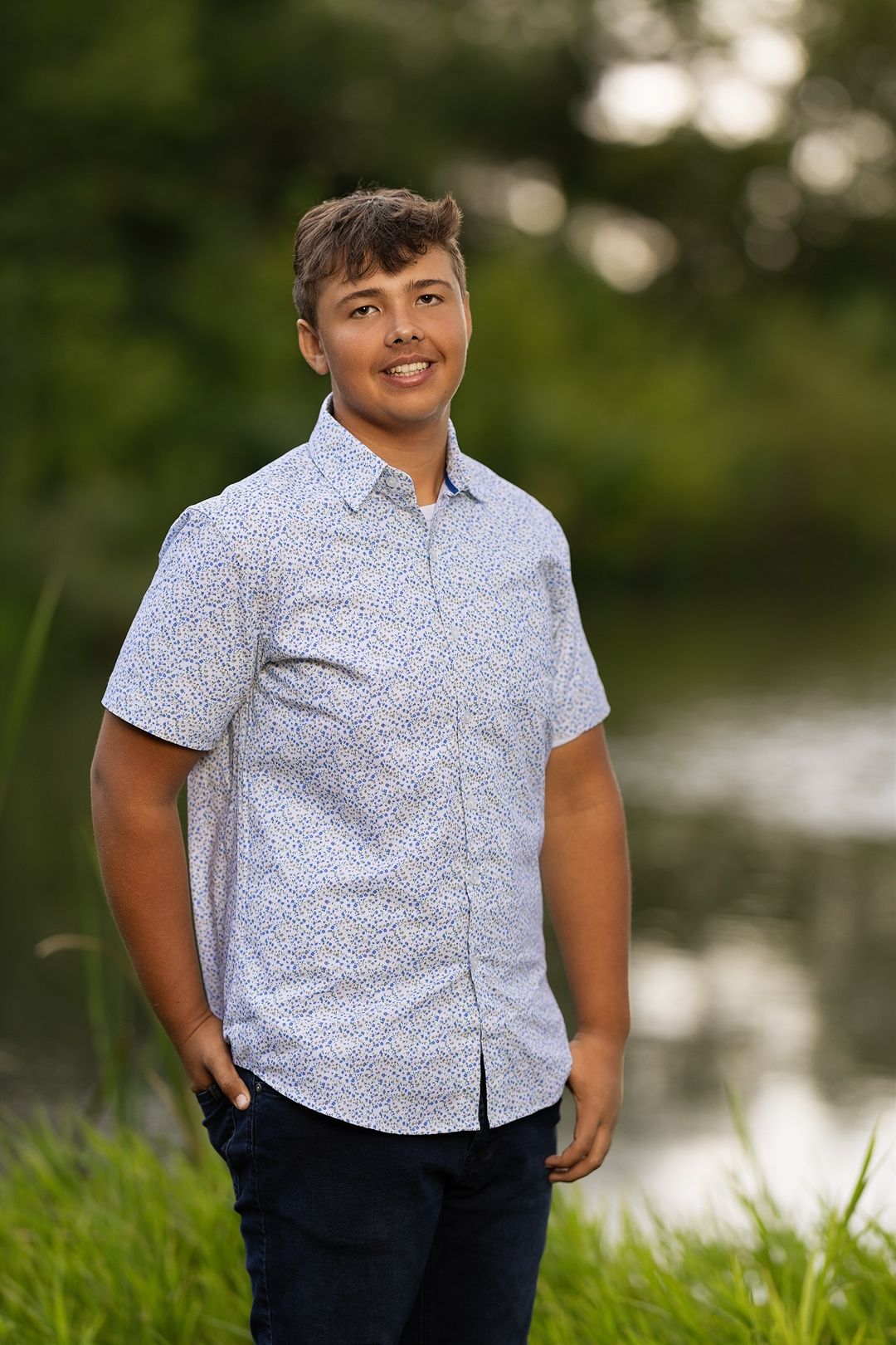 A person in a white patterned button-down shirt and dark pants poses outdoors near a lake in natural lighting.