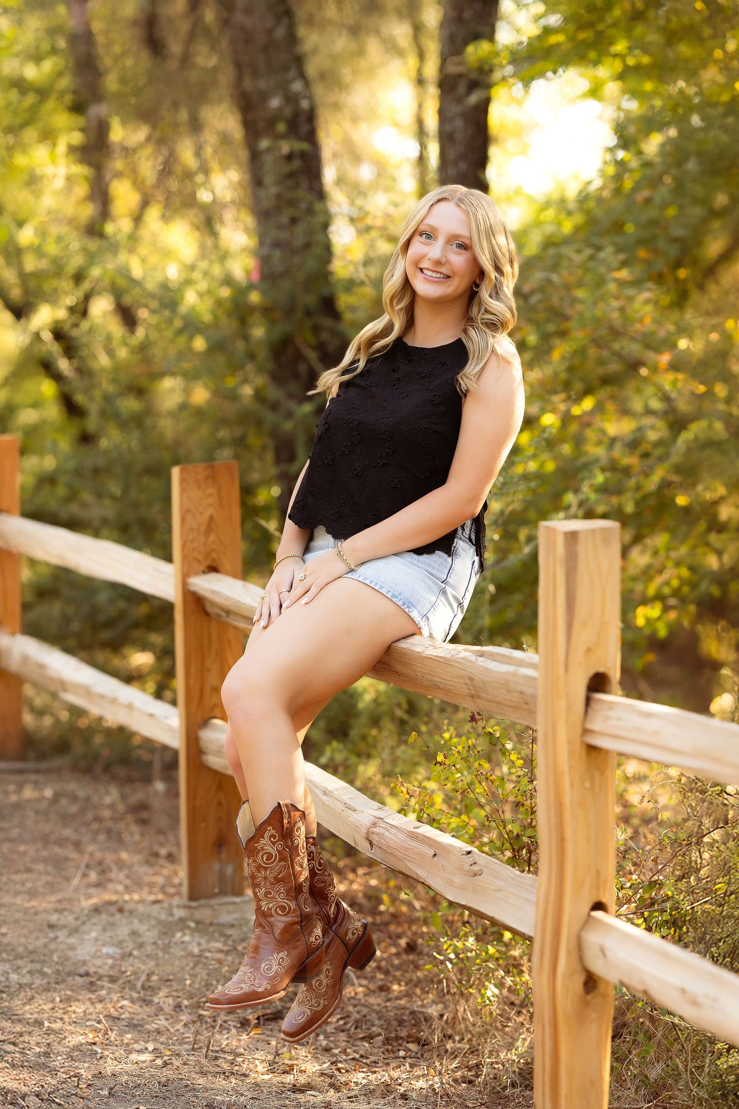 A person in cowboy boots sits casually on a wooden fence rail in a sunlit outdoor setting with autumn foliage.
