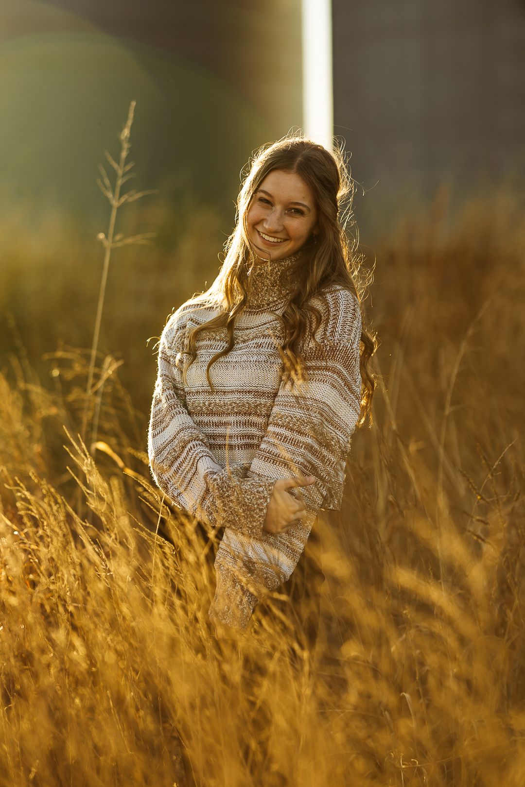 Person in cozy sweater laughs while sitting in golden sunlit field of tall grass at sunset.
