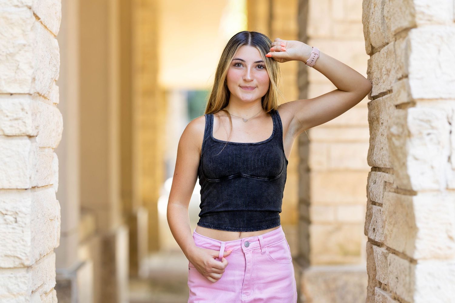 A person in a black top and pink pants poses confidently against a stone pillar in an outdoor setting.