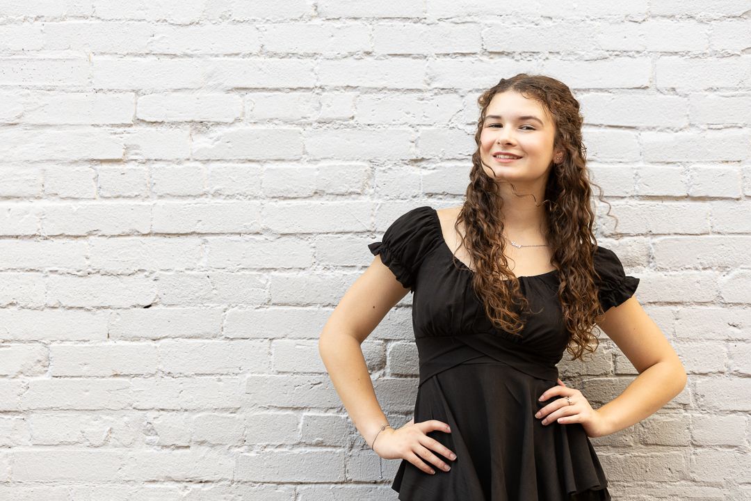 Woman in black dress poses playfully against white brick wall in photo series.