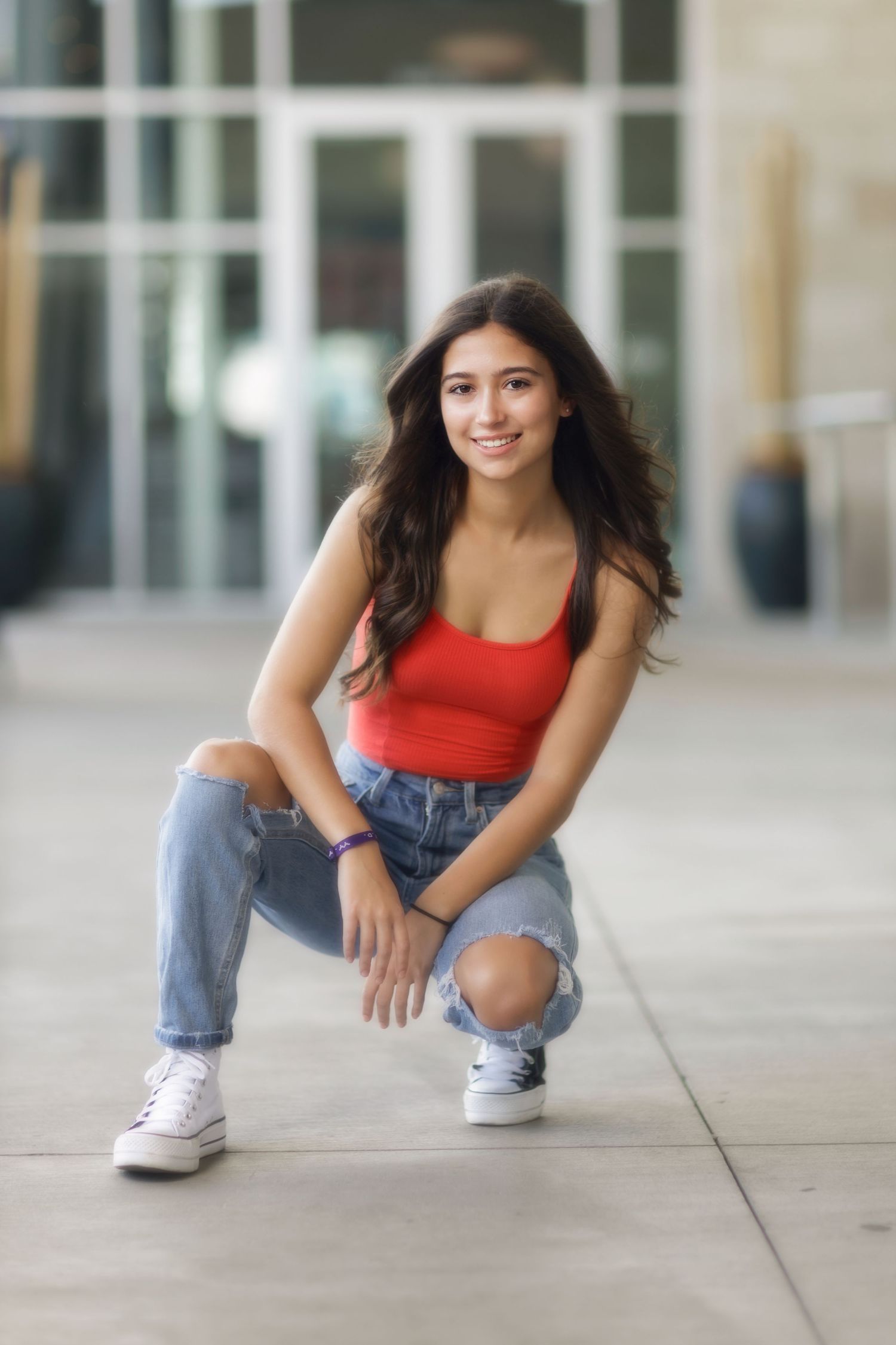 Young woman in red tank top and ripped jeans crouches on sidewalk wearing white sneakers in casual urban setting.