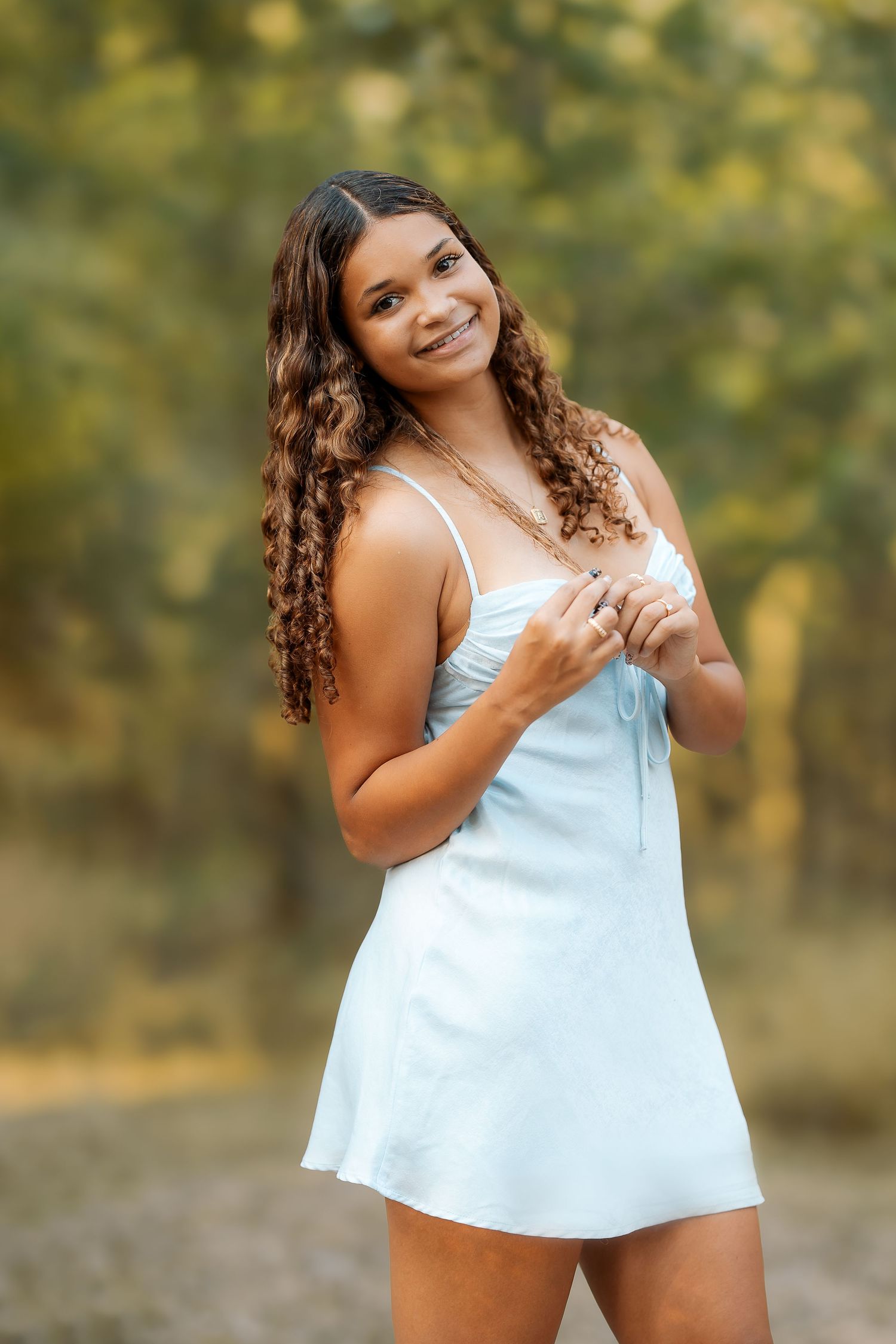 A woman in a white sundress poses outdoors with curly hair and natural lighting.
