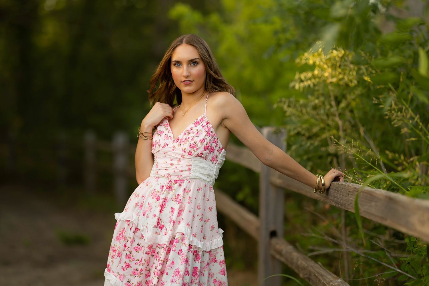 A person in a floral pink and white sundress poses outdoors near wooden fencing and lush green foliage.