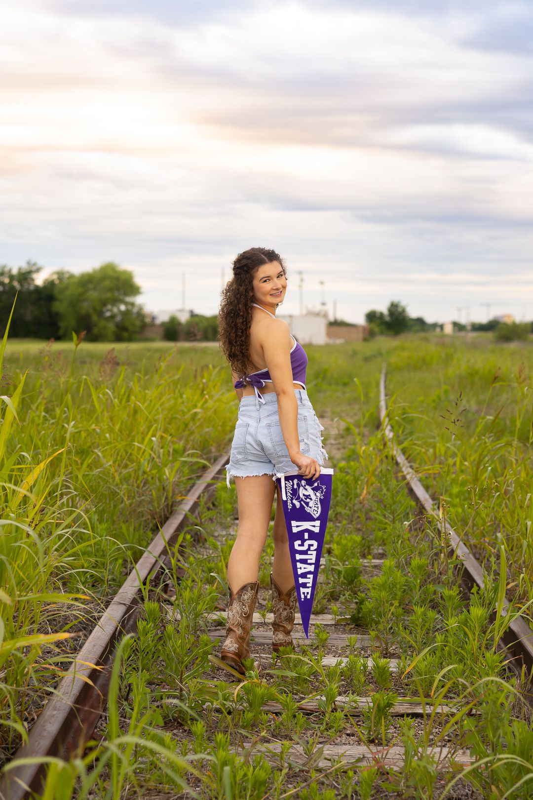 Old railroad tracks run through a grassy field with grain silos visible in the distance.