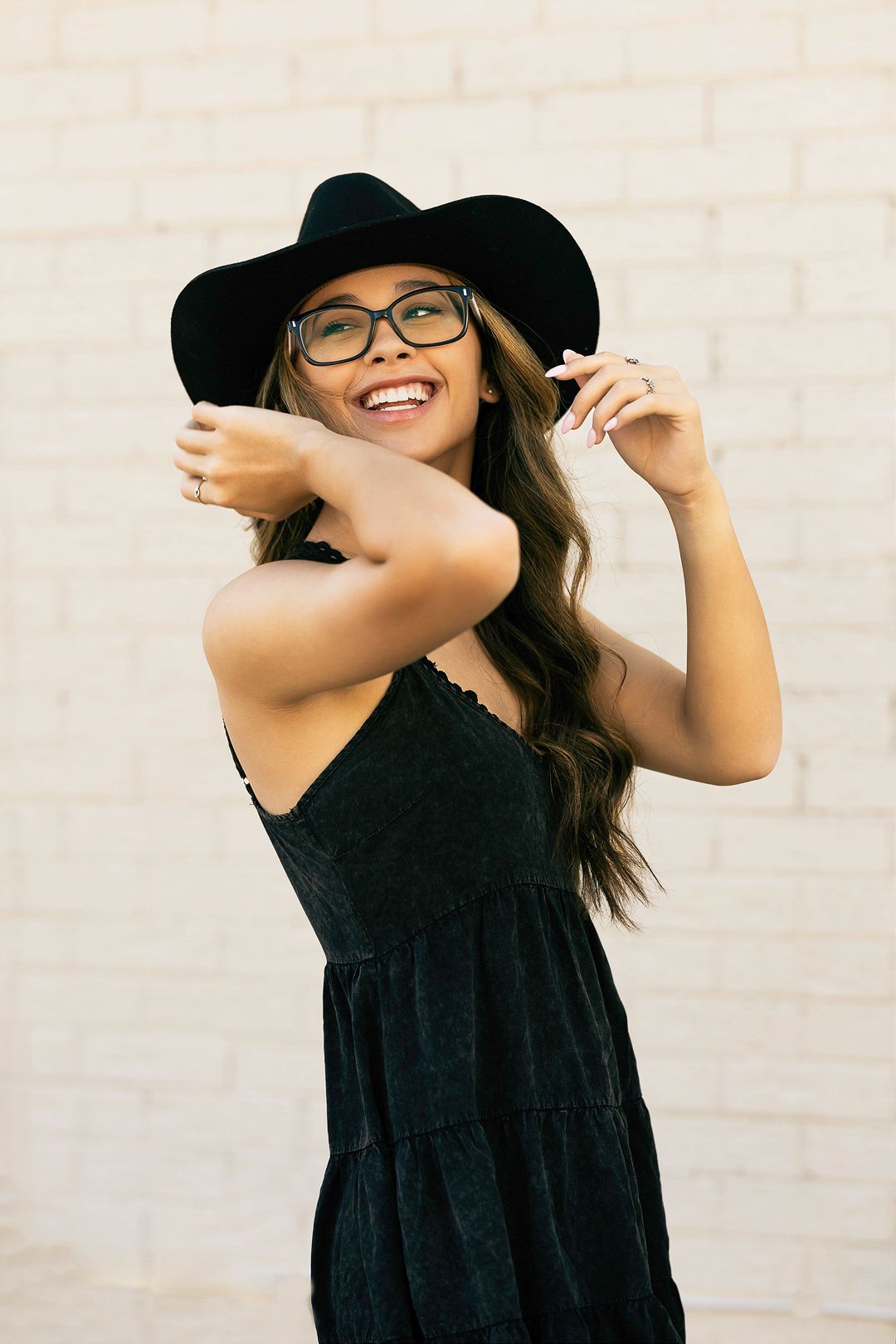 Playful poses in a black dress and wide-brimmed hat against a white wall.