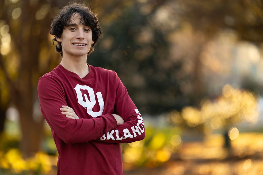 A person wearing a maroon Oklahoma sweatshirt stands outdoors in front of autumn foliage.