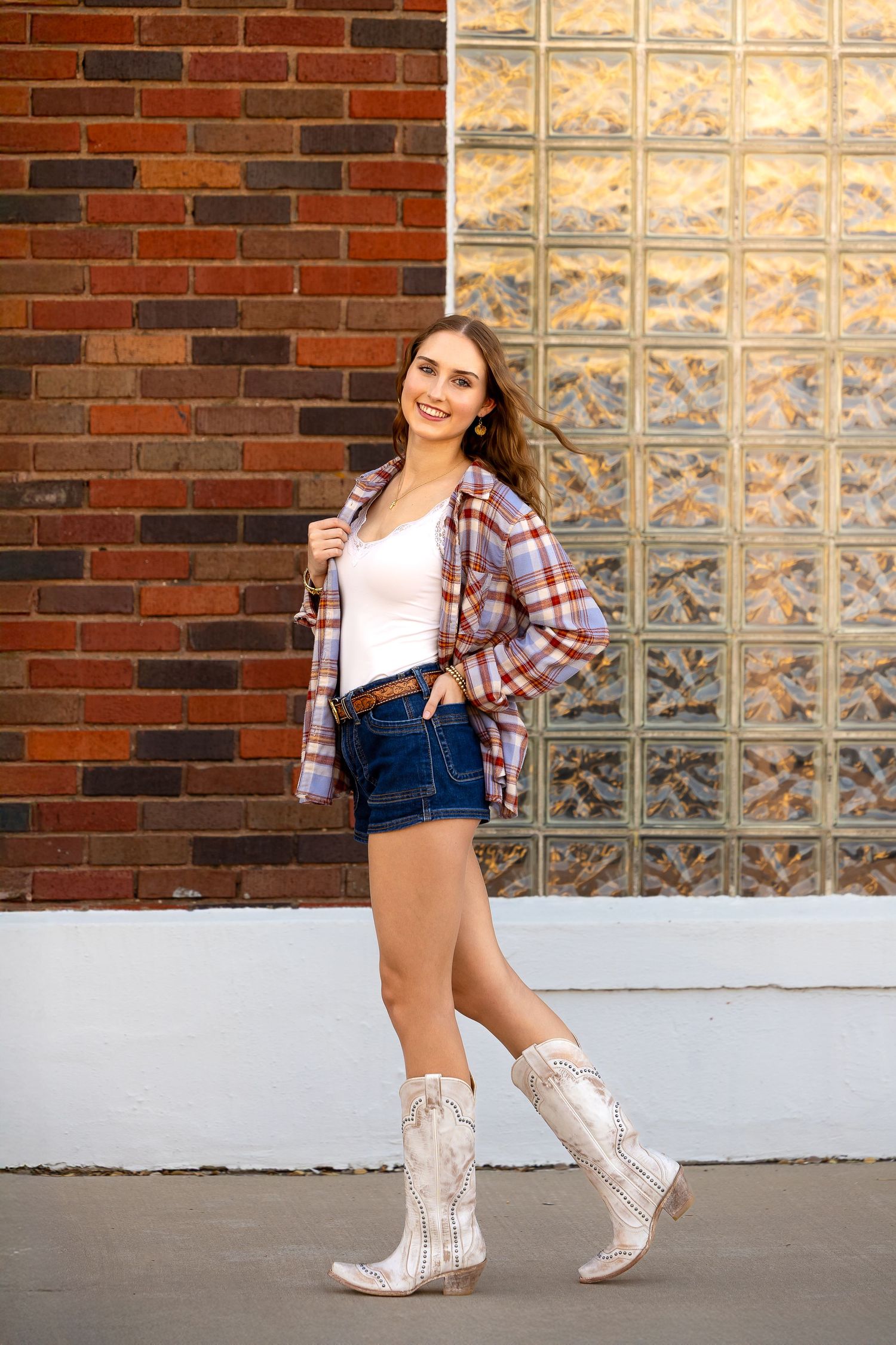 A person in denim overalls, plaid shirt and white cowboy boots poses against a brick wall with glass block windows.