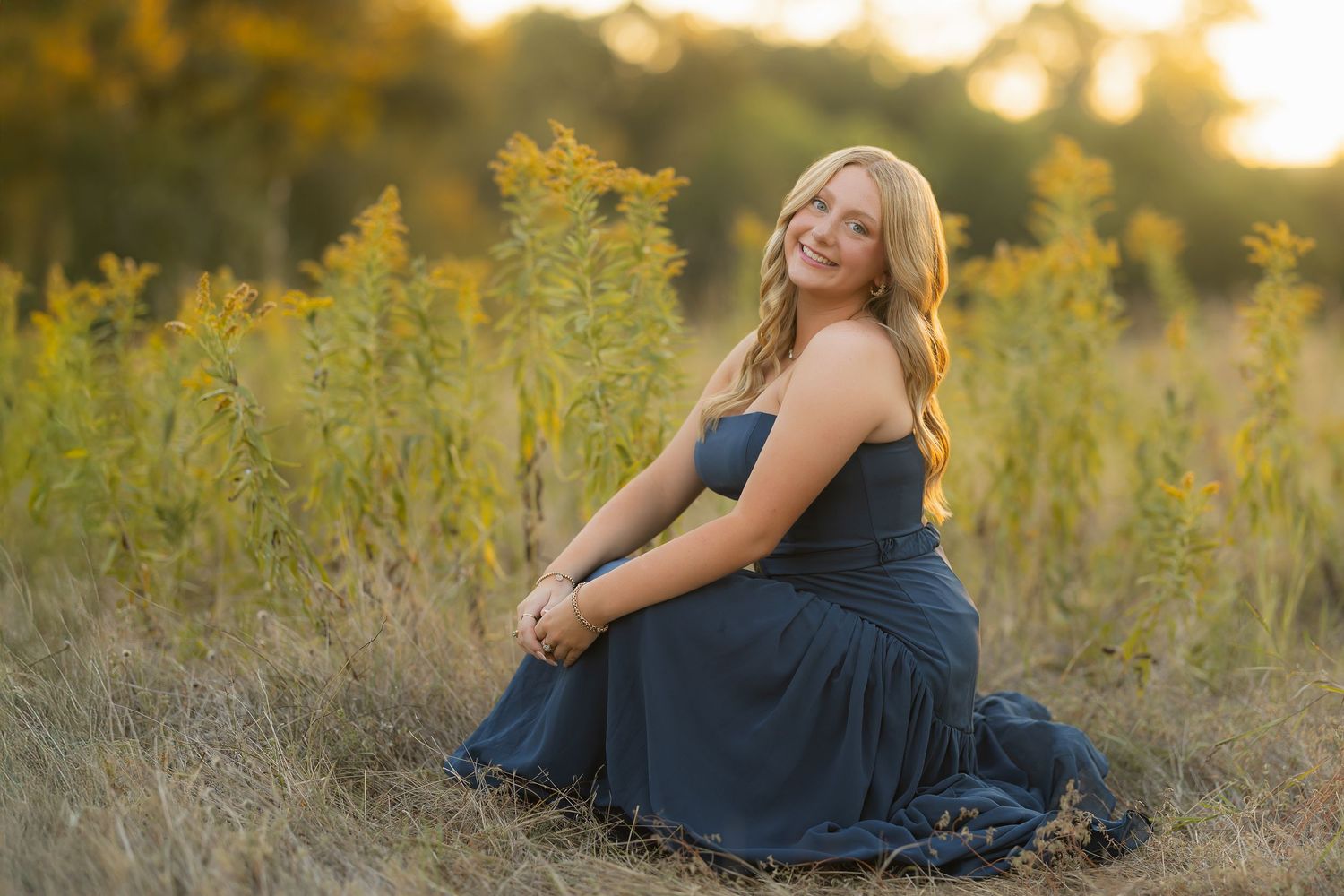 A person in a navy blue dress sits gracefully in a golden field during sunset creating a dreamy outdoor portrait.