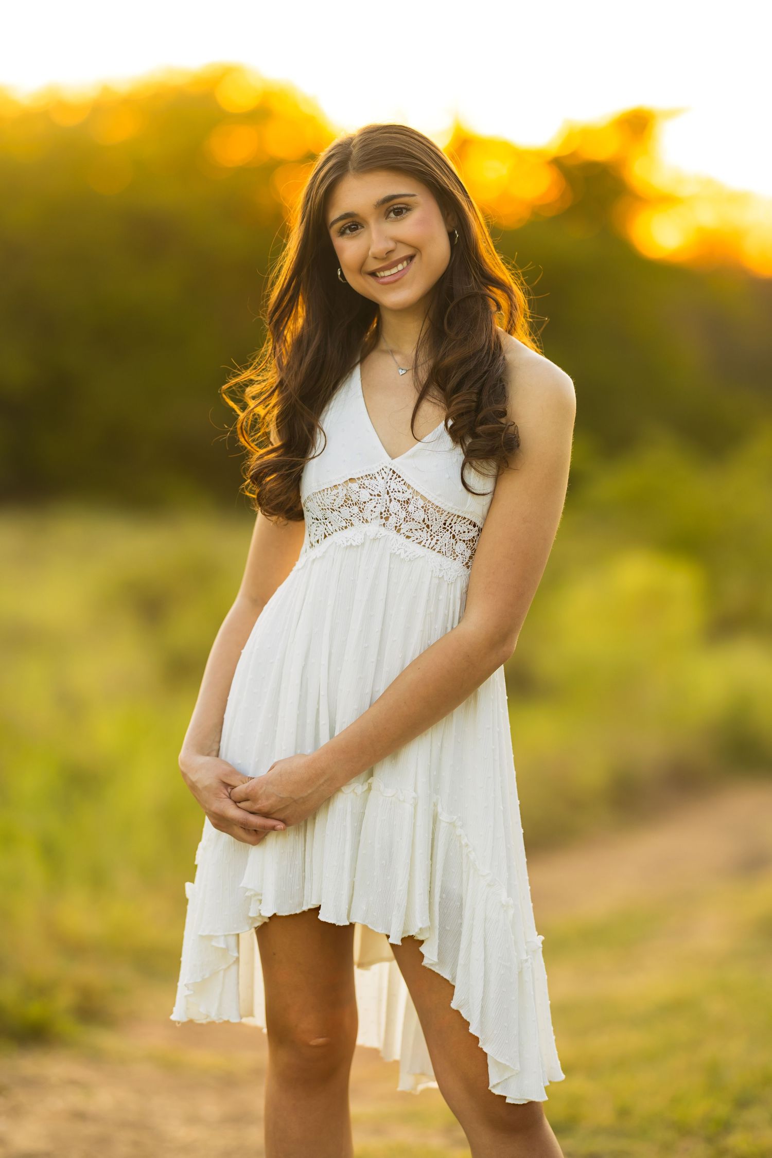 A woman in a flowing white dress stands in a golden field at sunset with warm backlighting creating a dreamy atmosphere.