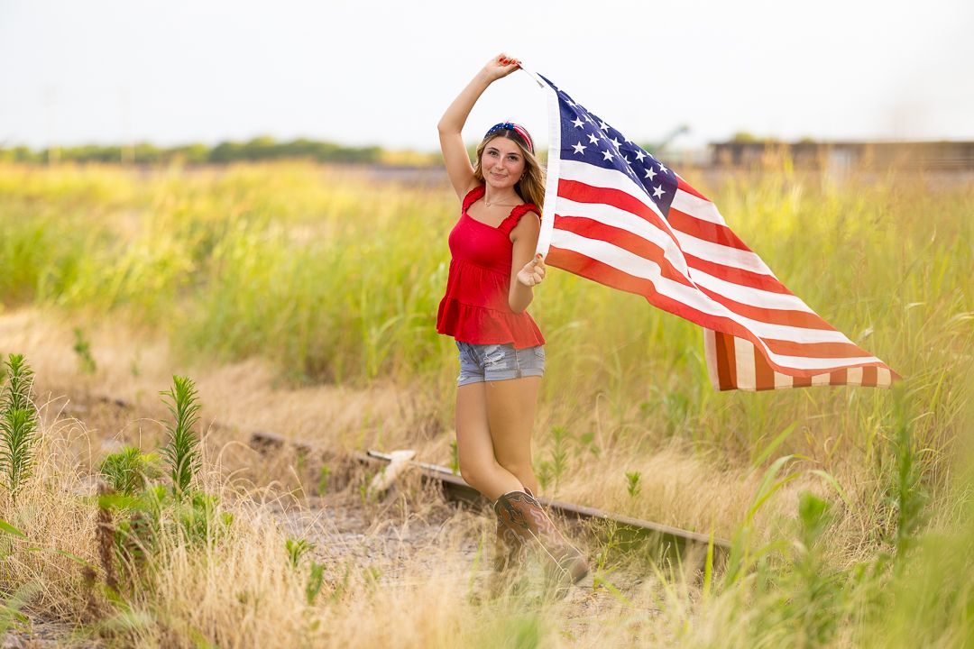 Young woman in red top and denim shorts waves American flag while standing on railroad tracks in a grassy field.