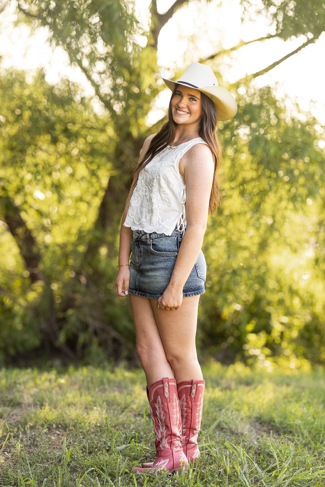 Smiling young woman in cowboy hat, white lace top, denim shorts, and red cowboy boots standing outdoors.