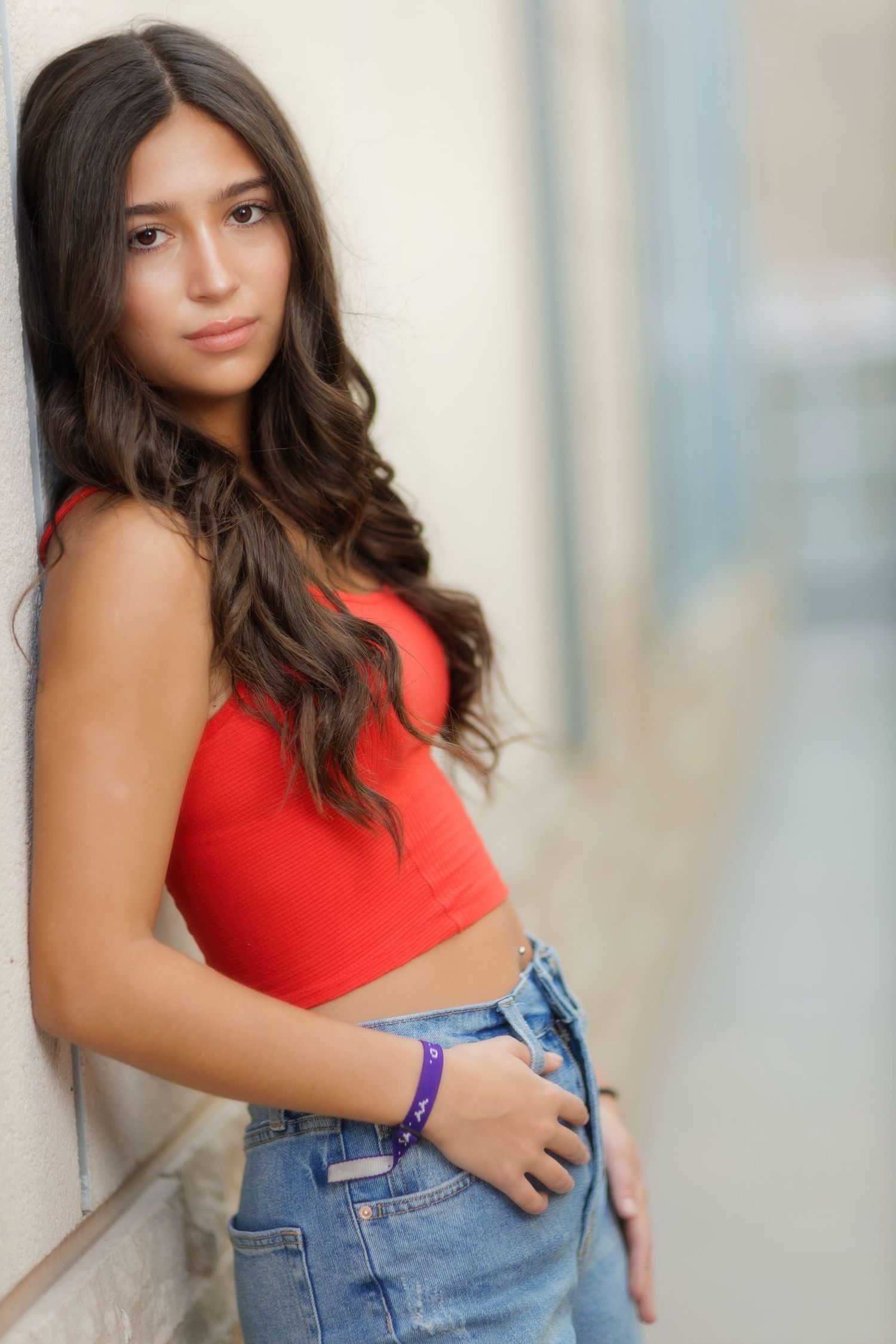A person in a red tank top and denim overalls poses against a light-colored wall in a casual photoshoot.