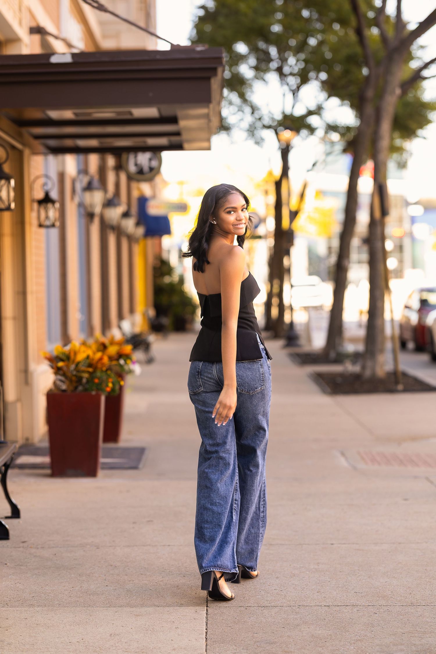 Person wearing blue jeans and black top walking along a covered outdoor shopping area with planters and trees.