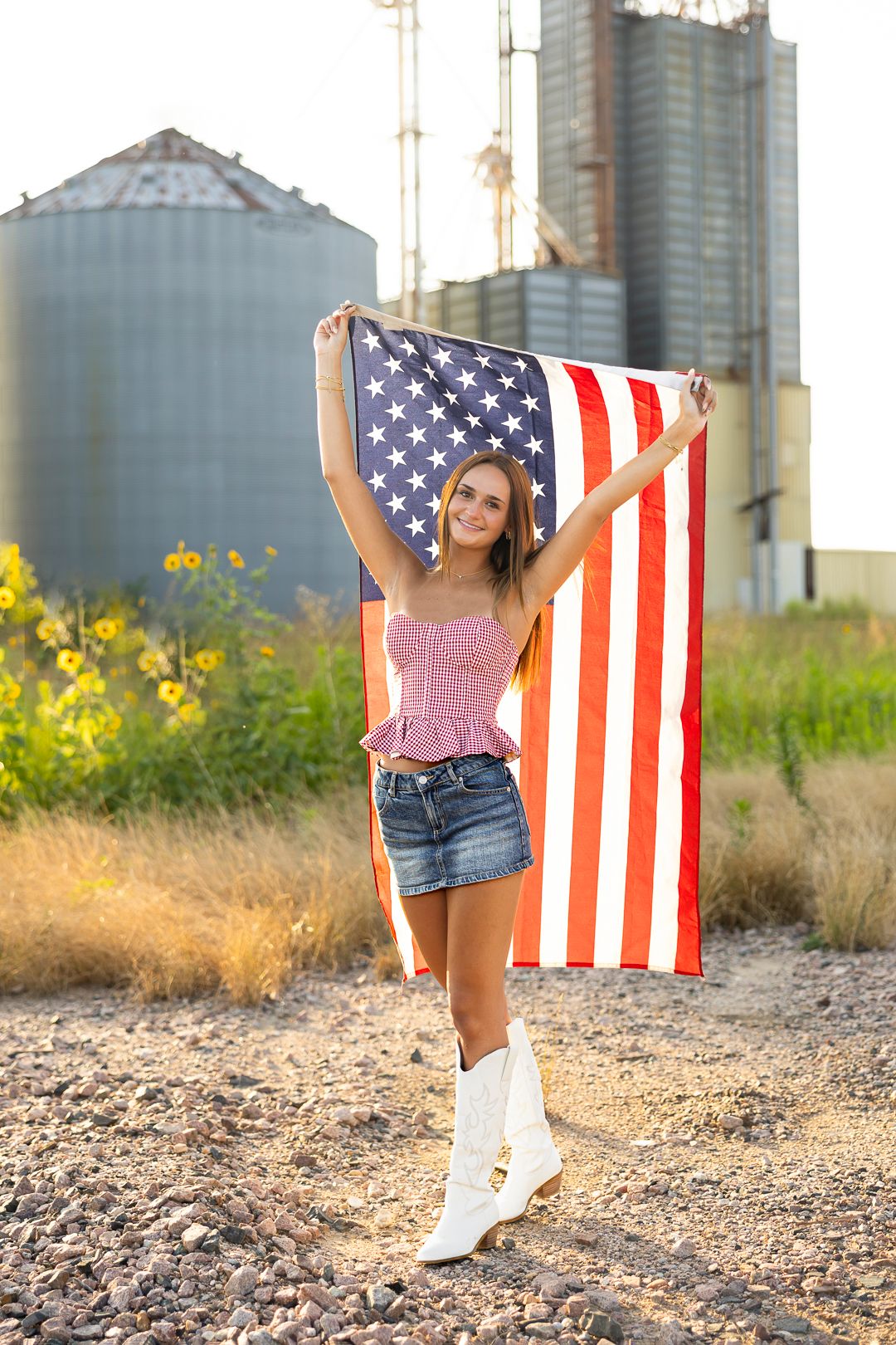 Young woman in denim skirt and cowboy boots holds American flag outdoors near grain silos on a sunny day.