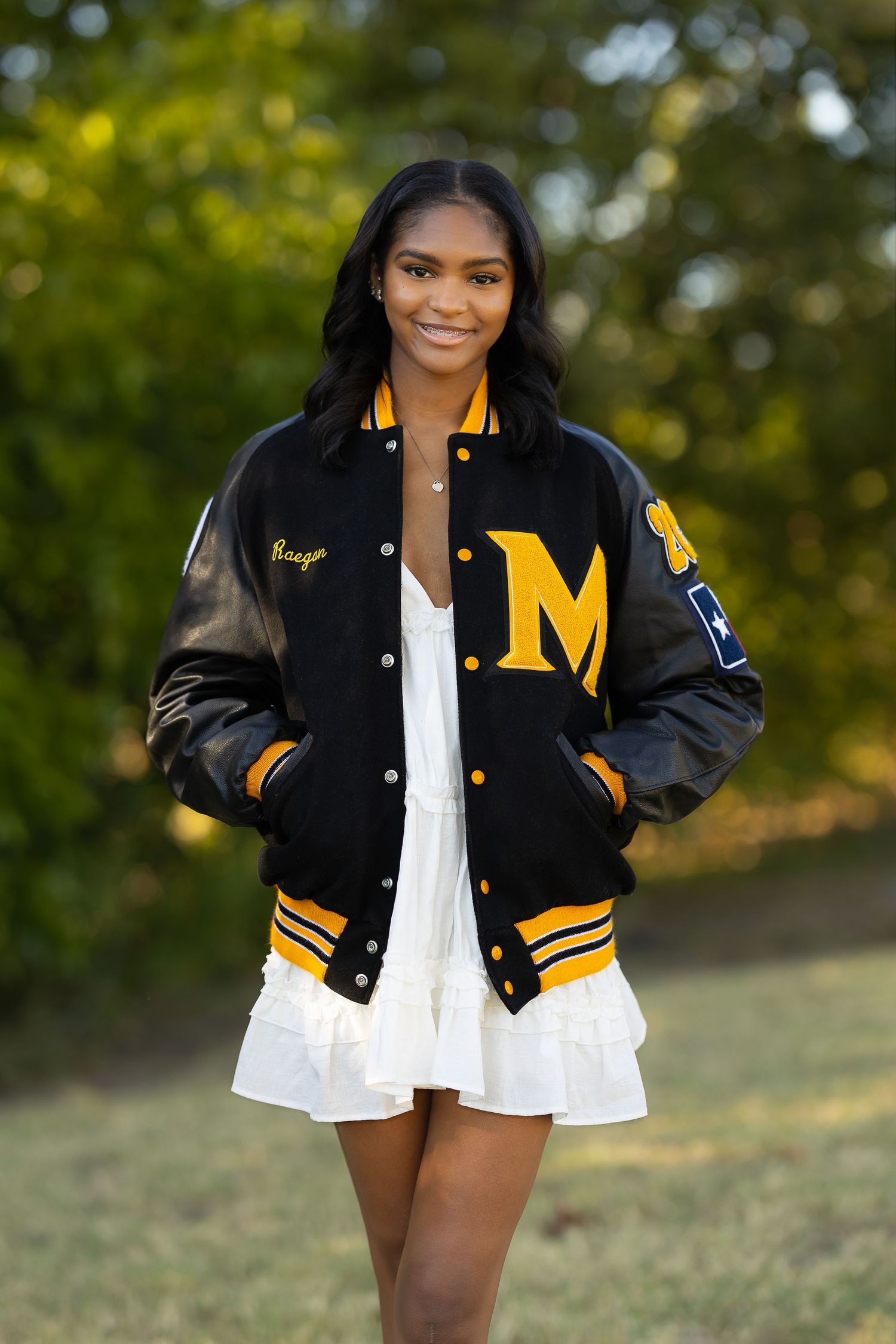 A varsity letterman jacket in black and gold paired with a white dress creates a stylish contrast in an outdoor setting.