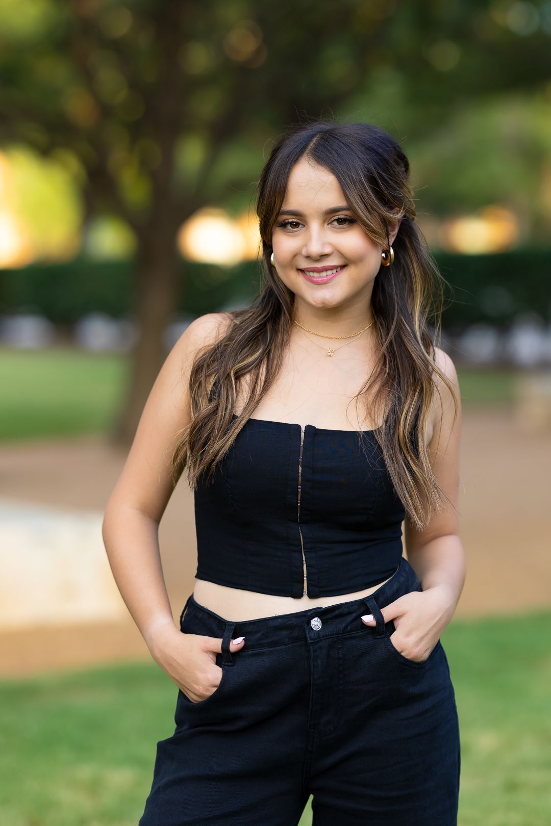 A person in a black outfit poses outdoors in a park with blurred green foliage in the background.