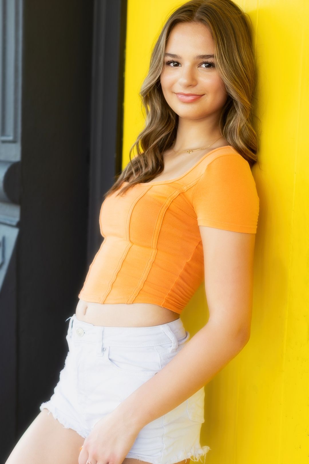 Young woman in orange top and white skirt poses against bright yellow wall in outdoor portrait session.