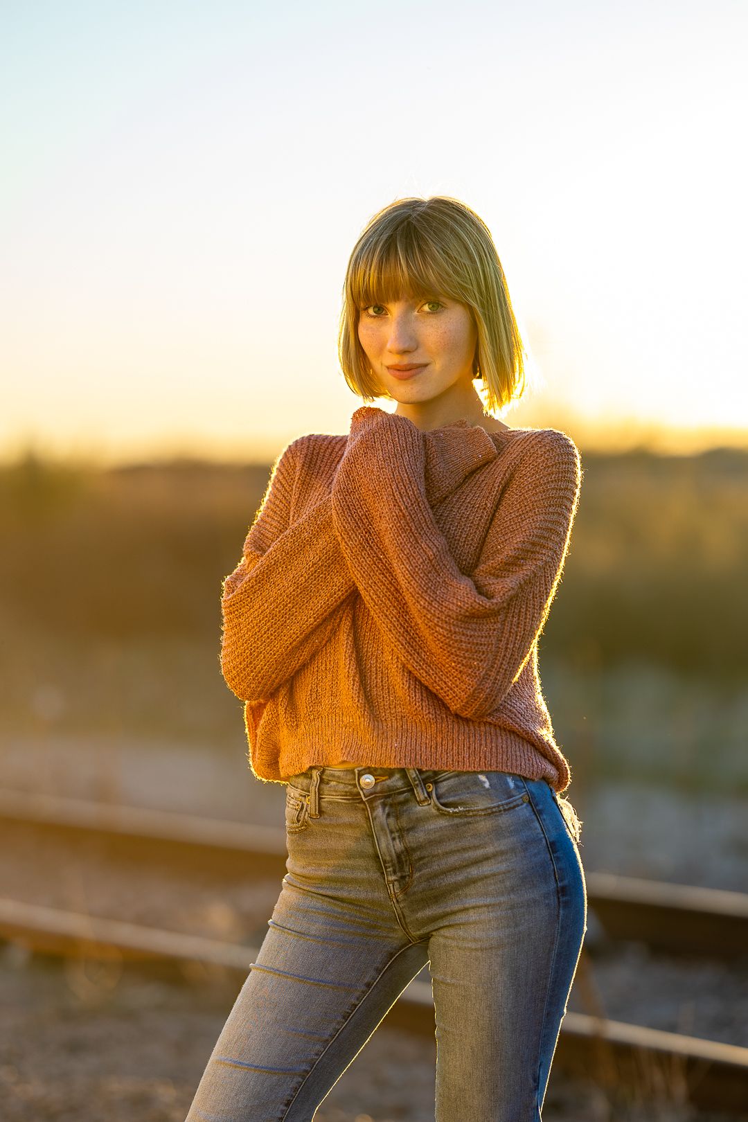 Woman in orange sweater and jeans poses against golden sunset light on rooftop.