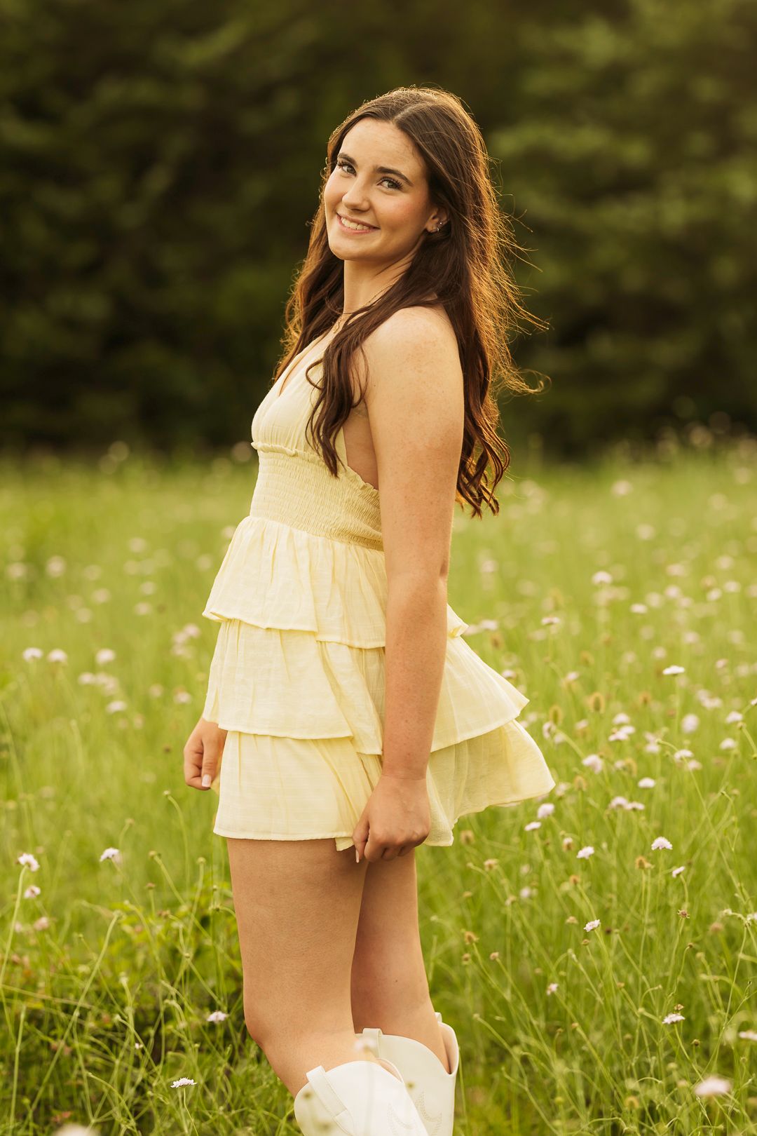 Young woman in yellow ruffled mini dress and white boots smiling in a wildflower meadow.
