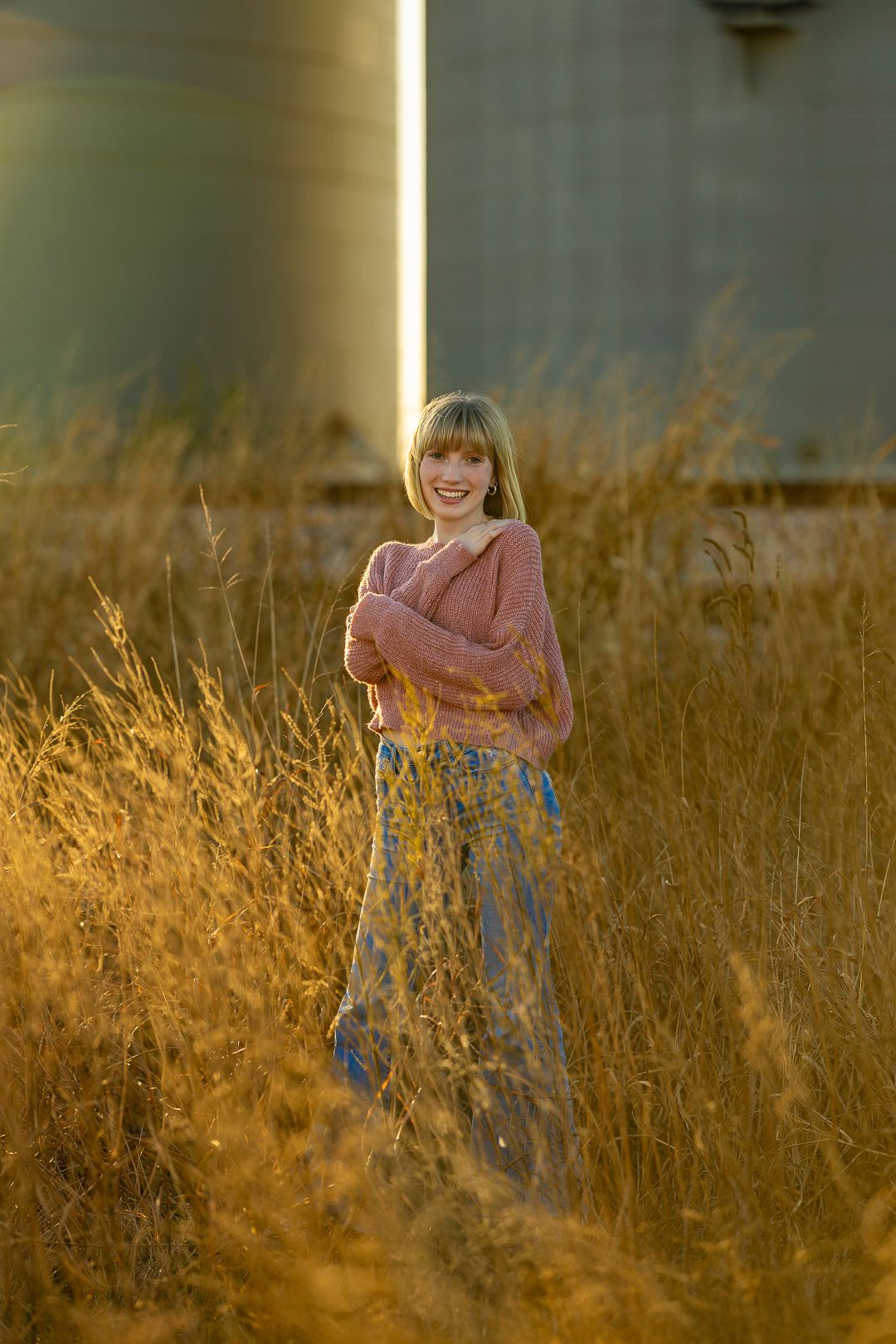 Figure in pink sweater stands among tall golden grass during sunset.