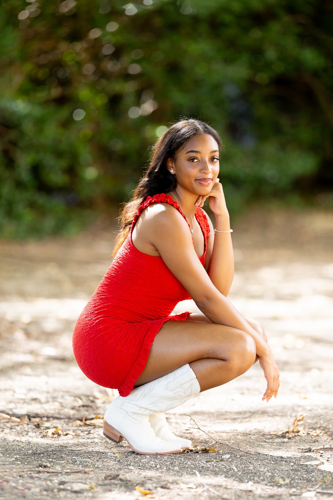 A person in a red sleeveless top and white shorts crouches on a gravel path in a scenic outdoor setting.