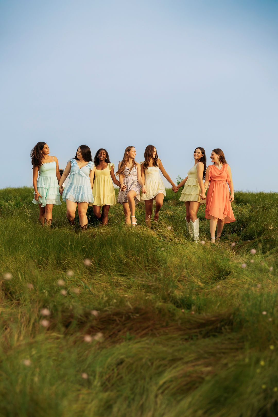 Seven young women in colorful pastel dresses walk together through a lush green meadow under a clear blue sky.