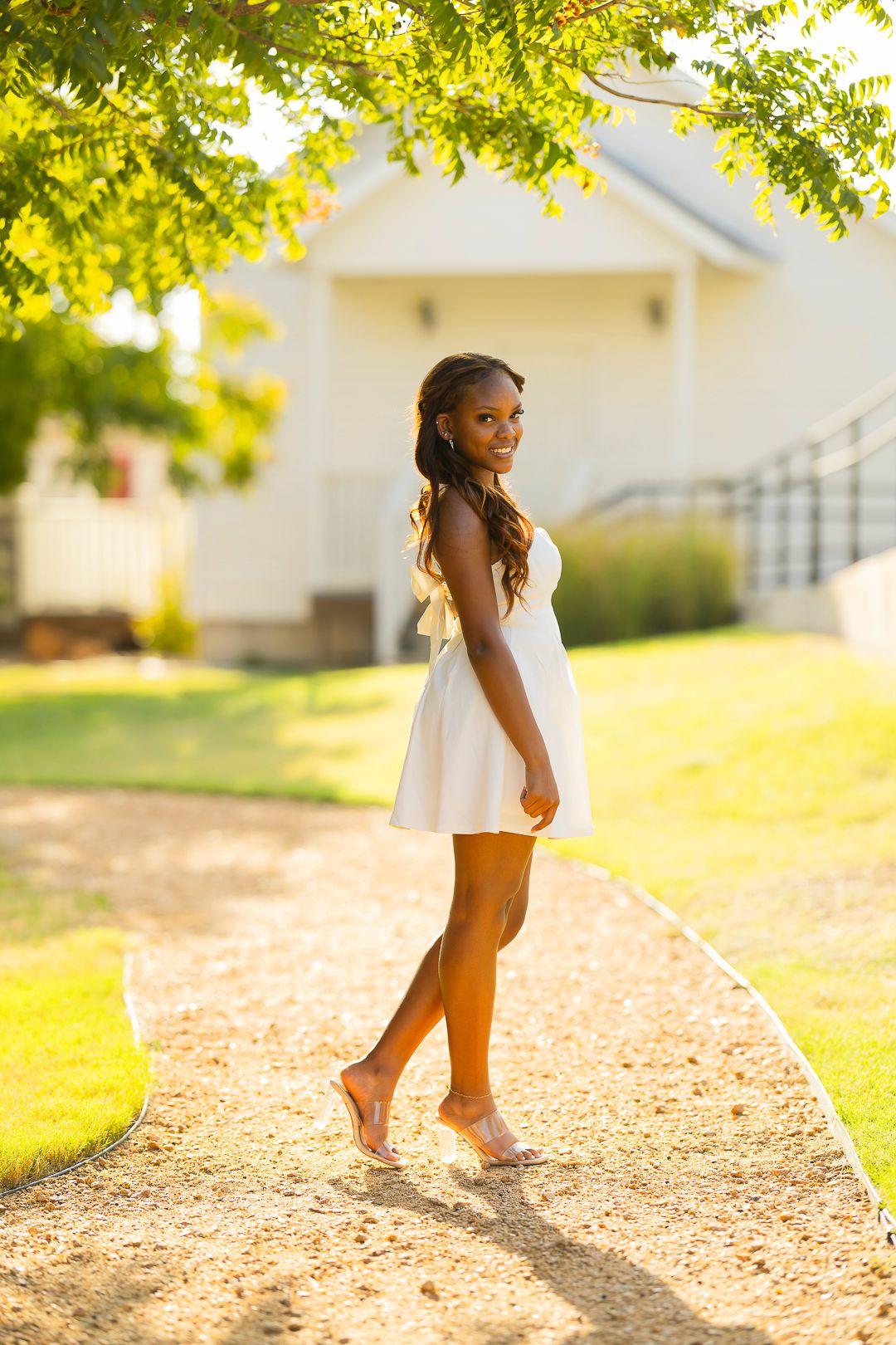 A person in a white dress strolls along a sunlit gravel path under trees.